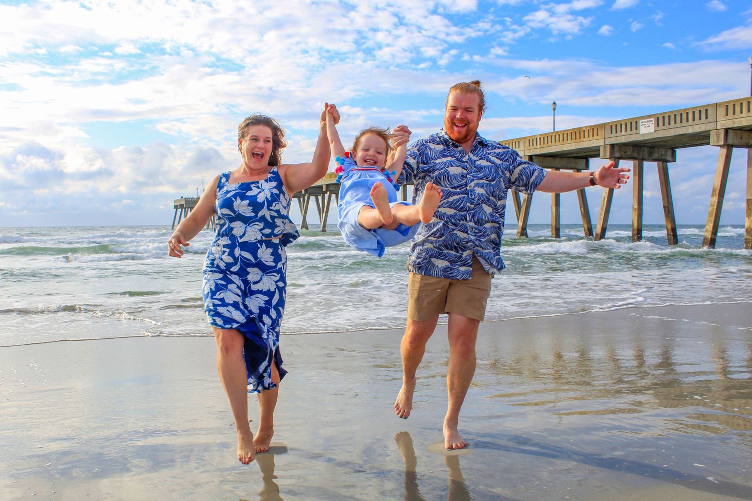 A joyful family plays together on the beach near a fishing pier on a sunny day.