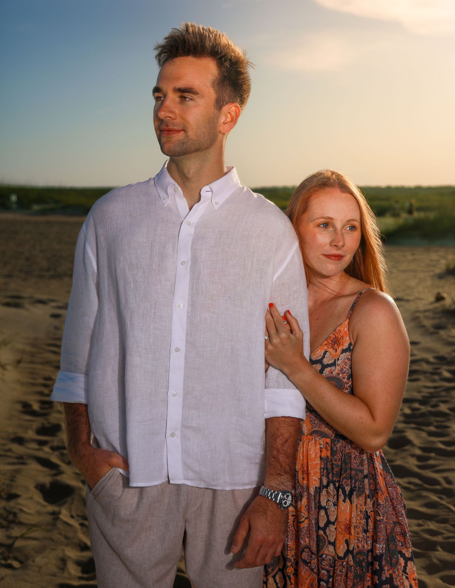Couple posing on sandy beach at sunset, man in white linen shirt, woman in floral dress leaning on his shoulder.