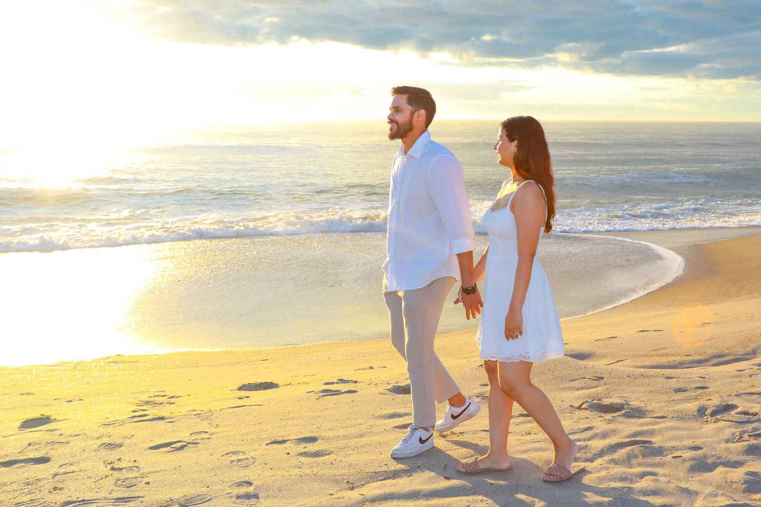 Couple holding hands walking on a sandy beach at sunset, wearing white outfits with ocean waves in the background.