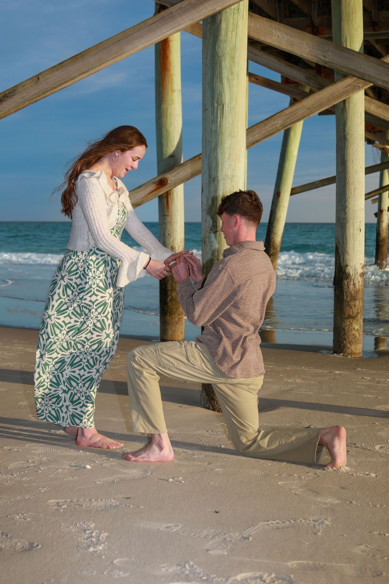 Man proposes on one knee to woman on sandy beach under pier with ocean waves in background.