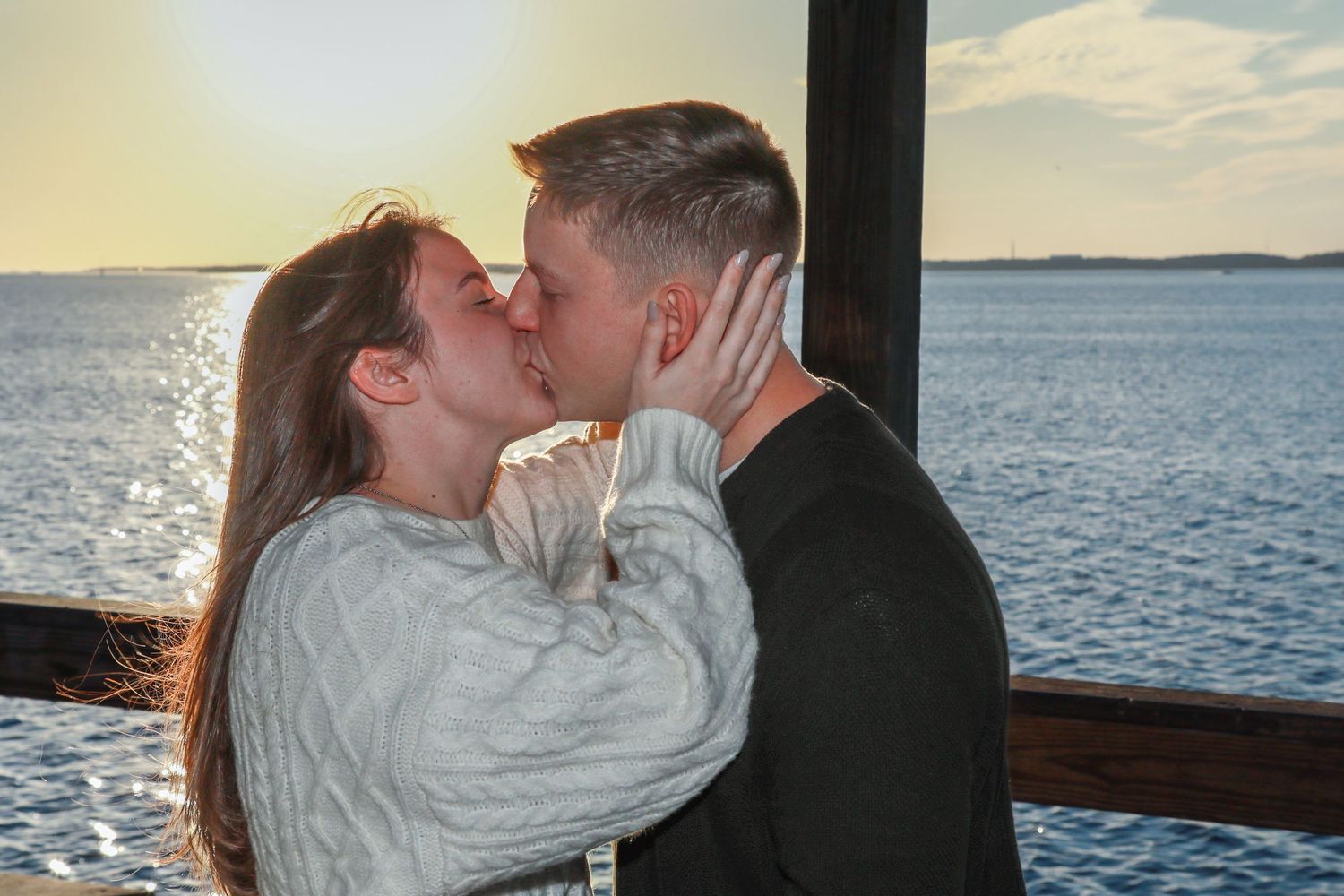 Couple sharing a romantic kiss at sunset by the ocean on a wooden pier.
