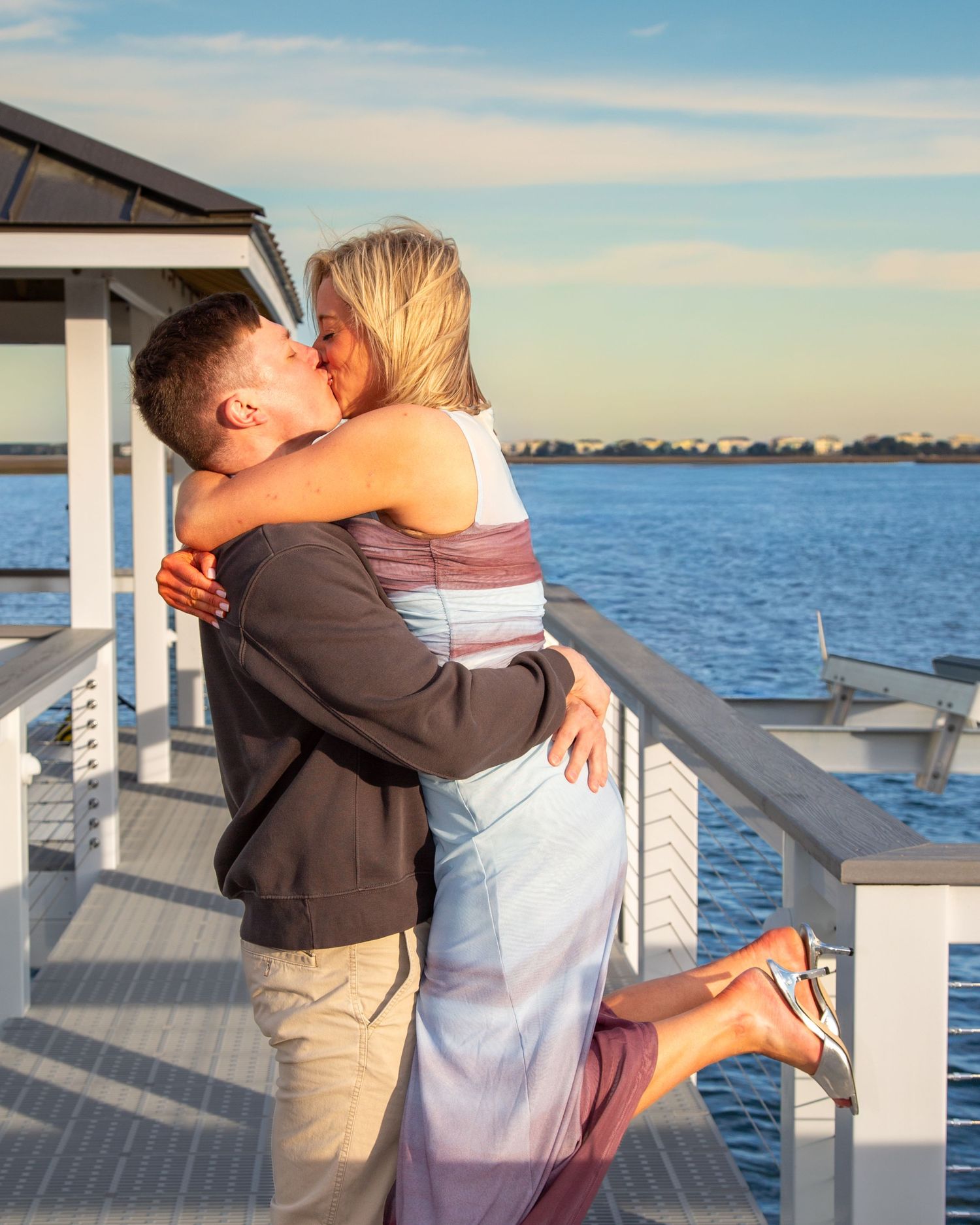 Couple sharing a romantic kiss on a waterfront deck at sunset, woman in colorful maxi dress lifted by man in gray jacket.