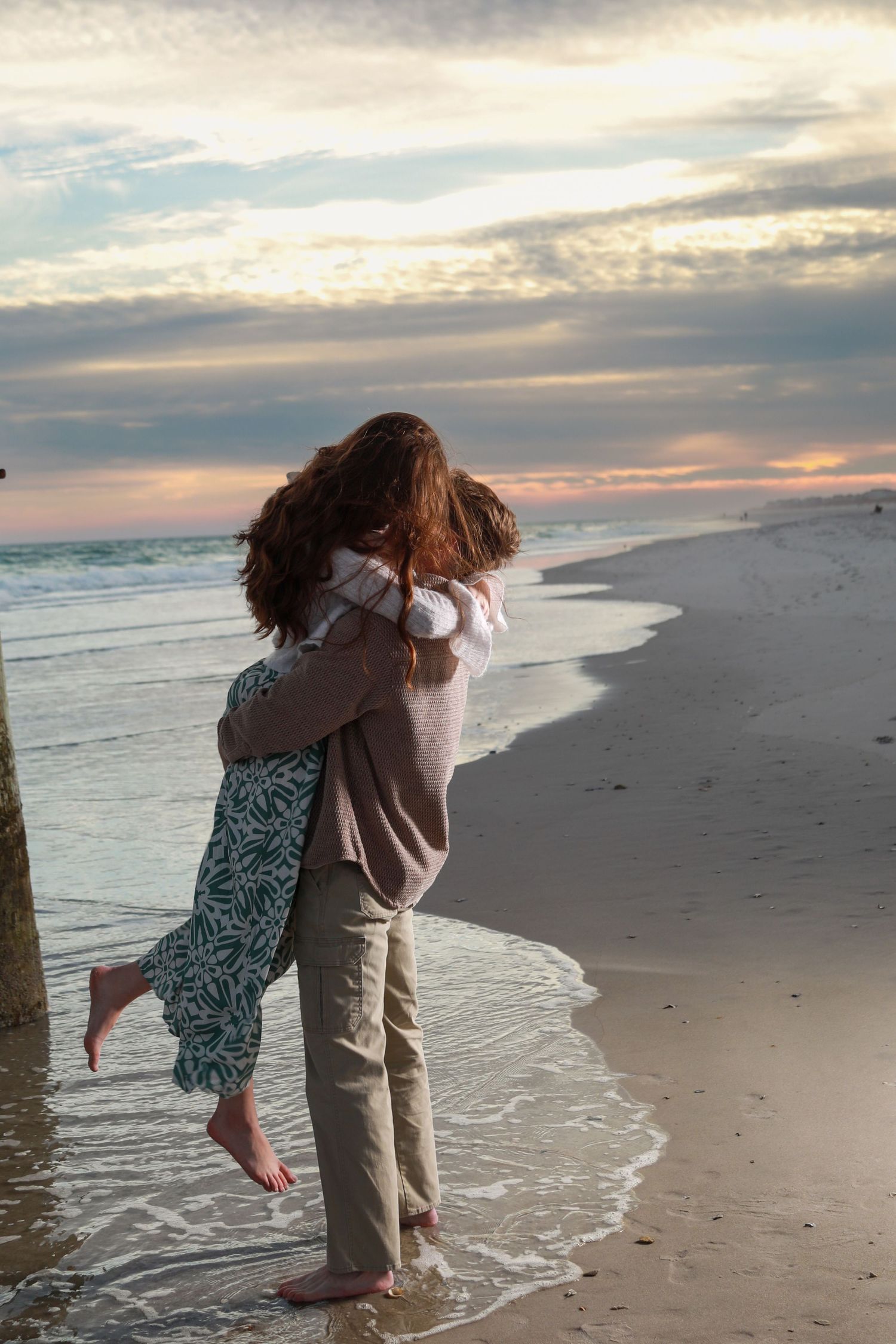 Couple embracing on a sandy beach at sunset, waves washing ashore under a colorful cloudy sky.