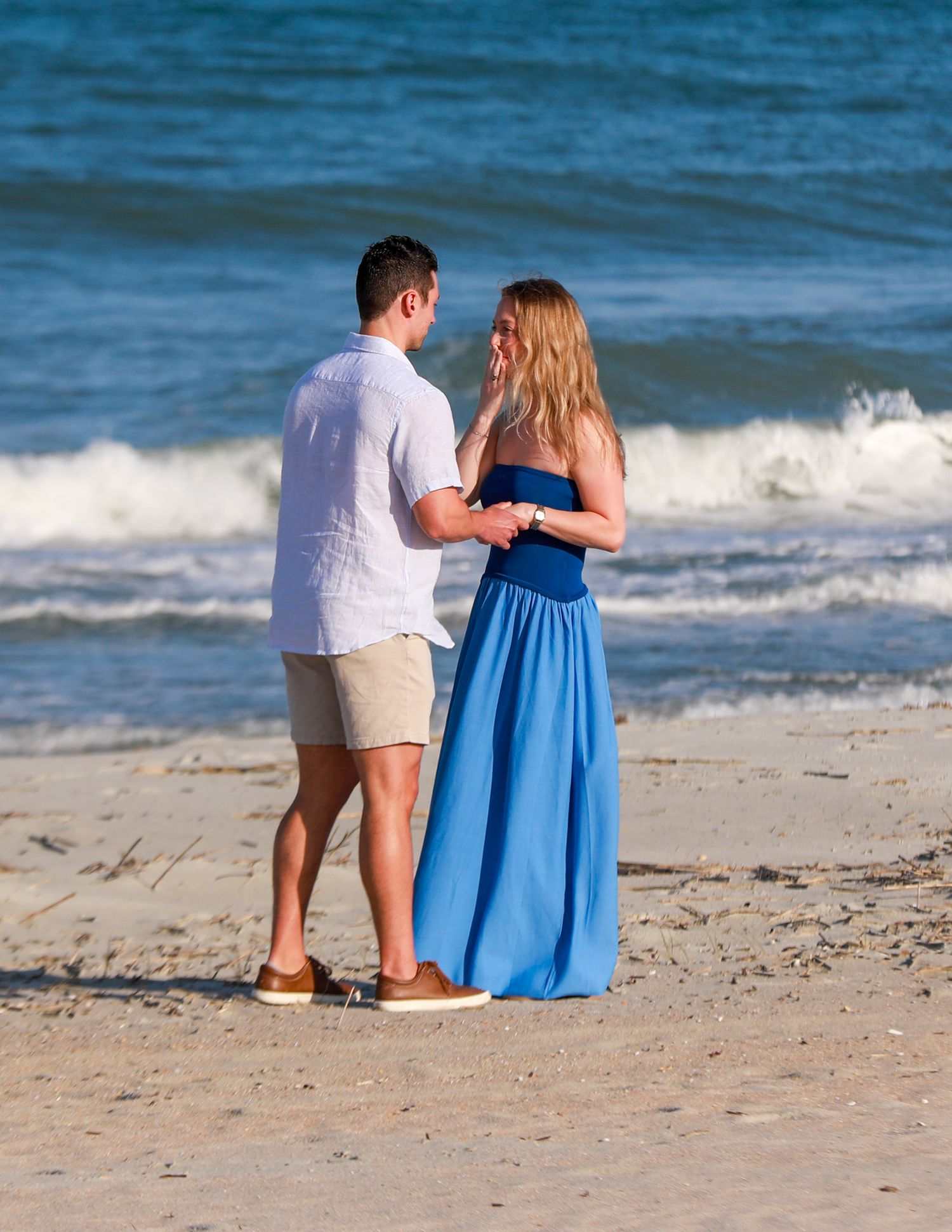 Couple holding hands on a sandy beach, woman in blue maxi dress, man in white shirt and khaki shorts, ocean waves behind.