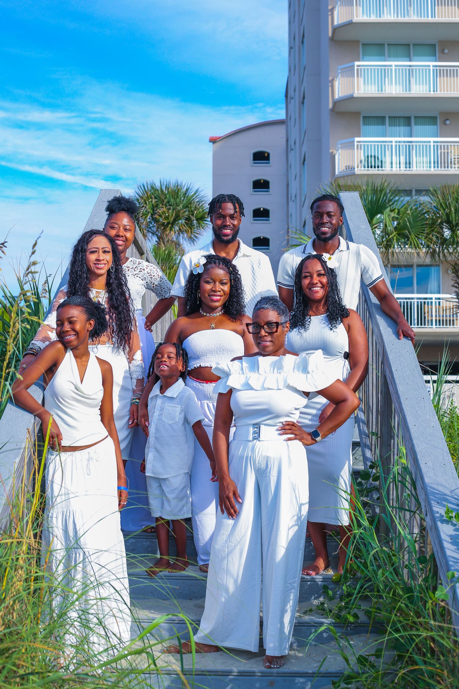 A joyful Black family poses in matching white outfits on a sunny beach boardwalk with palm trees and a hotel behind them.