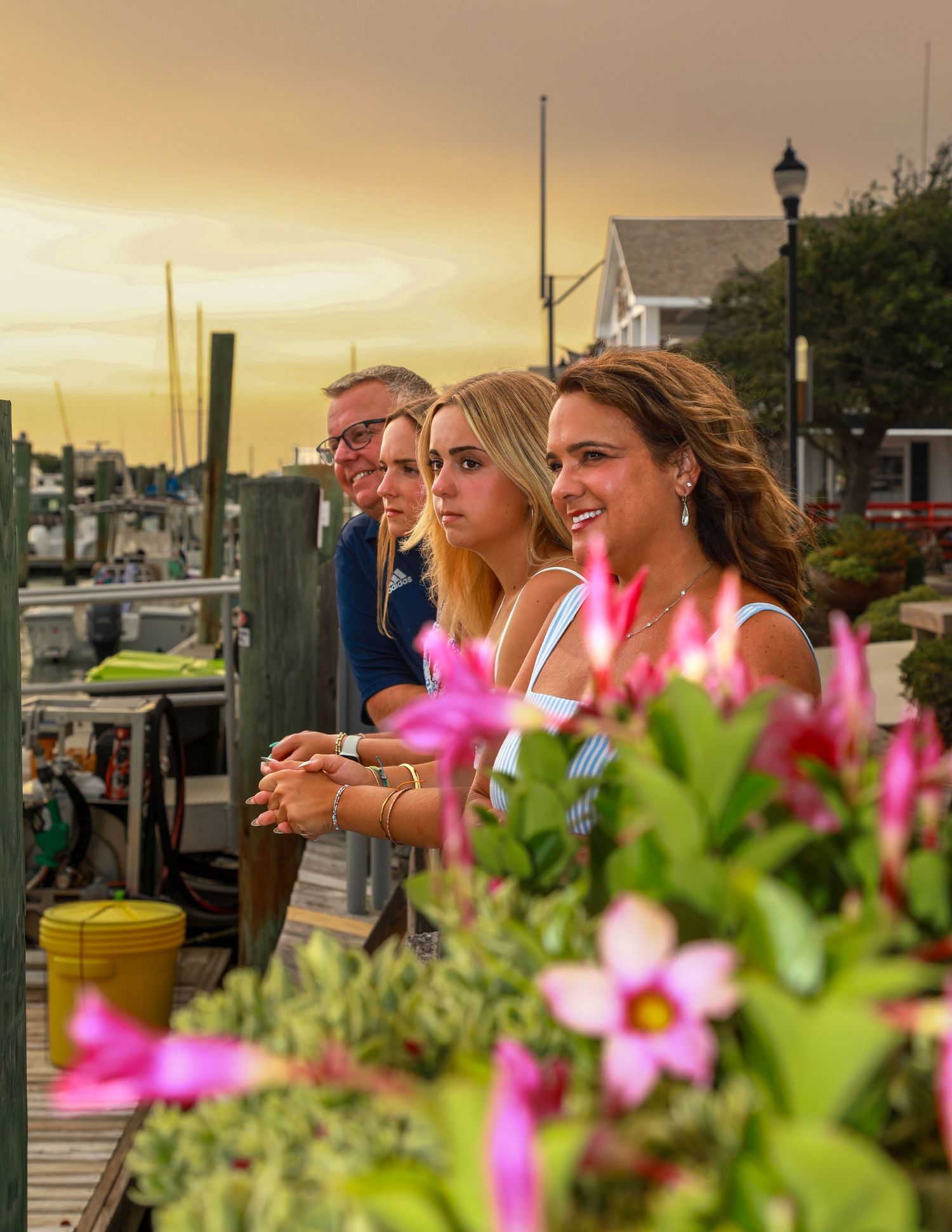 Family enjoying a waterfront sunset view from a dock surrounded by pink flowers at a marina.