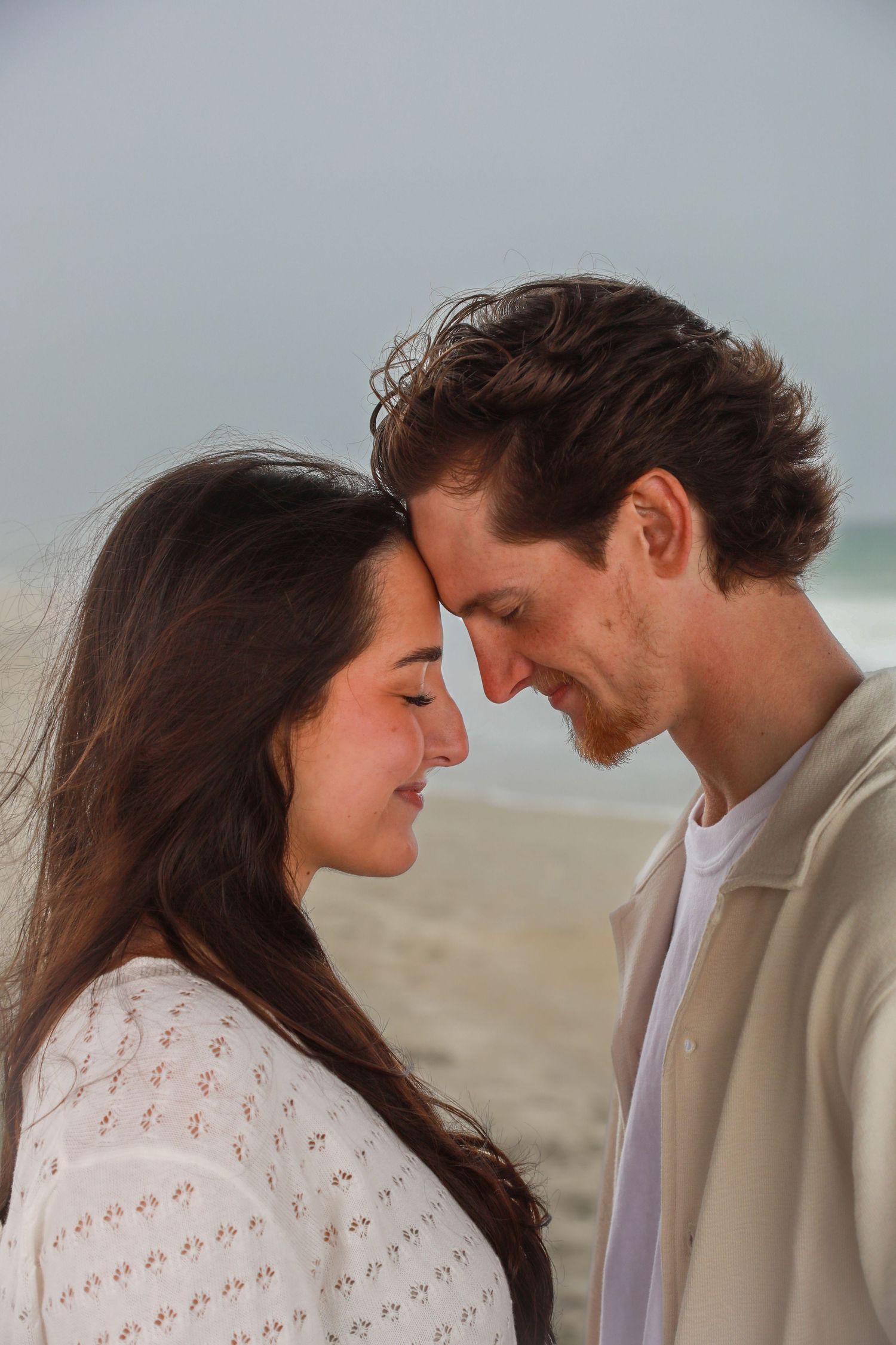 A romantic couple touches foreheads tenderly on a beach, sharing an intimate moment with ocean in background.