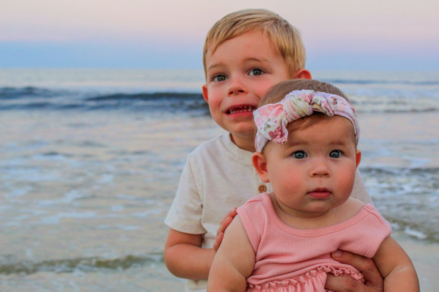 A baby in pink clothing sits on the beach during sunset.
