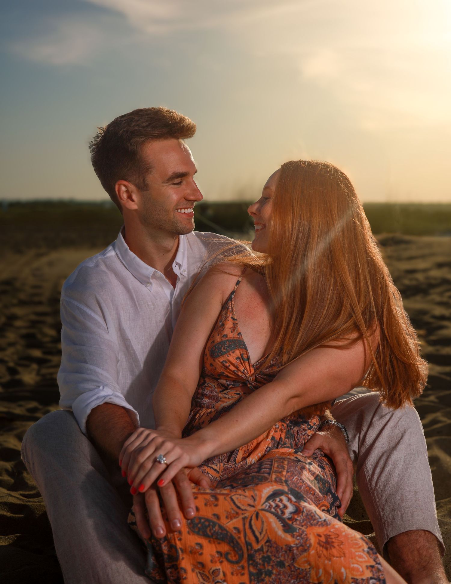 Romantic couple sitting on sandy beach at golden sunset, woman in floral dress smiling at man in white shirt.
