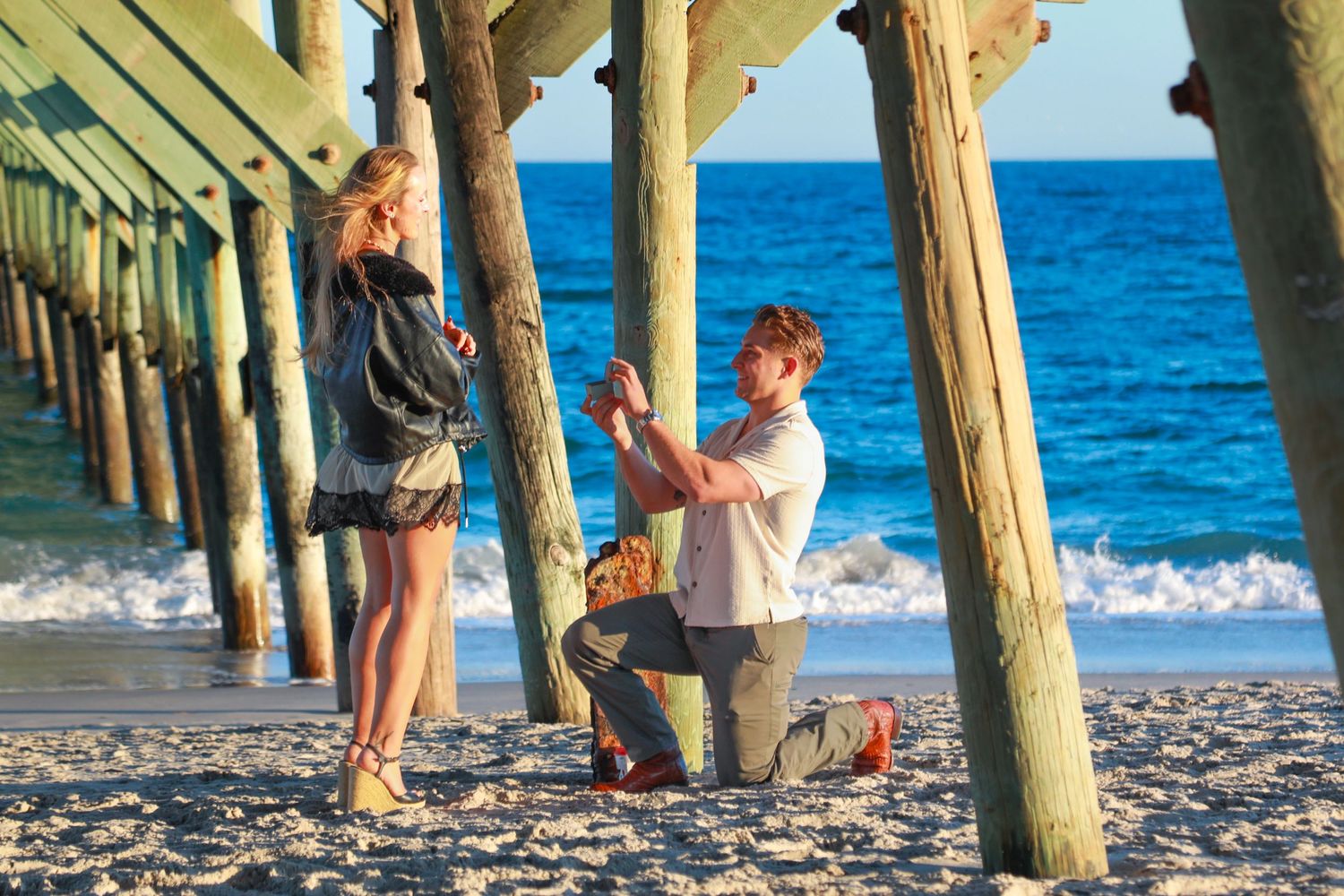 Man proposing on one knee to woman on sandy beach near wooden pier with ocean waves in background.