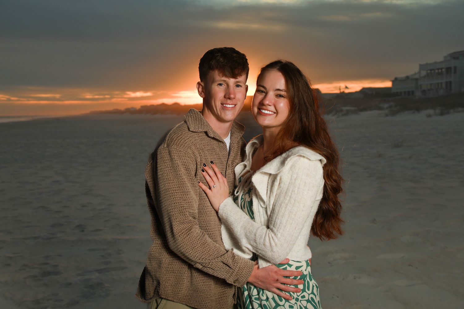 Couple embracing on a beach at sunset with golden sky and coastal buildings in the background.
