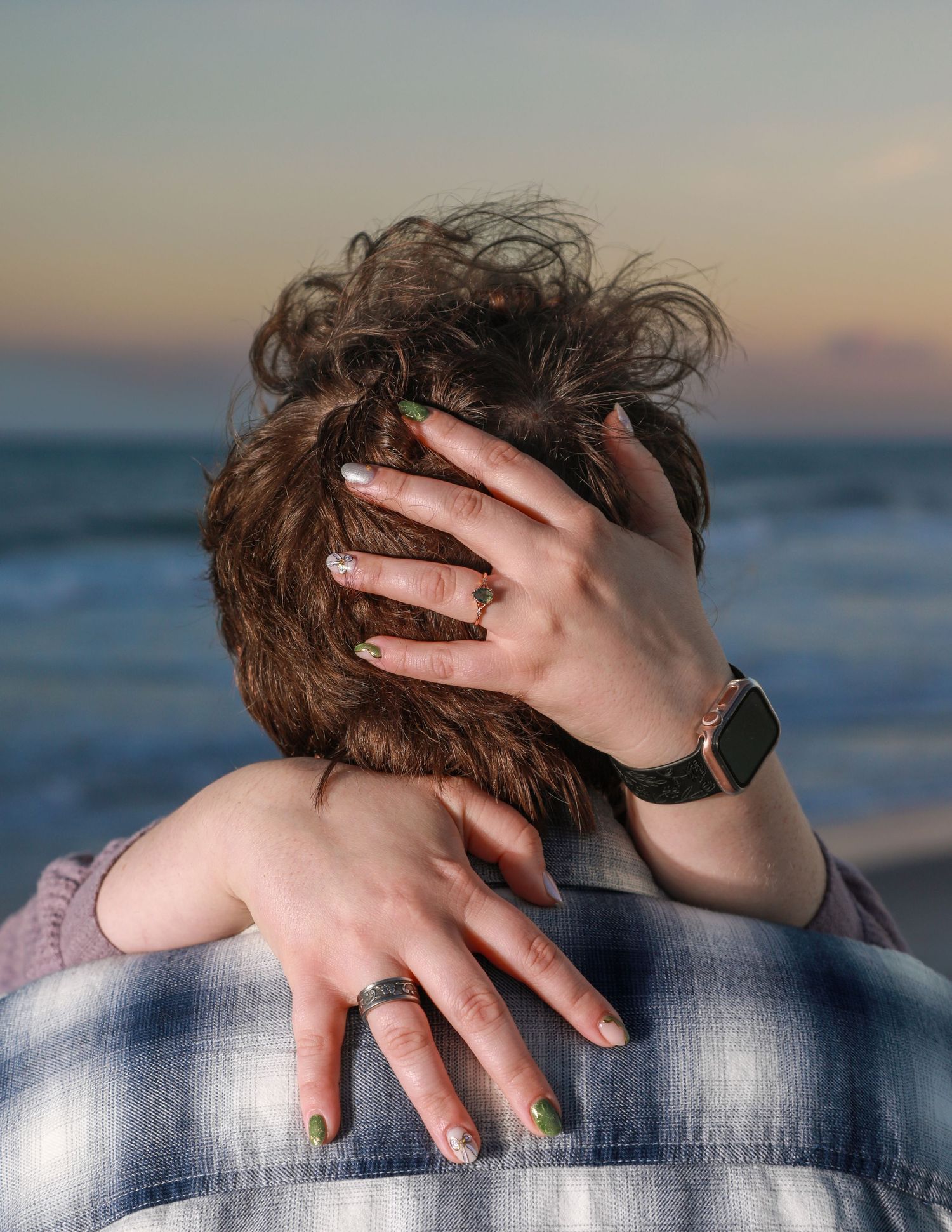 Couple embracing at sunset beach, woman's hand with ring and smartwatch resting on man's head.