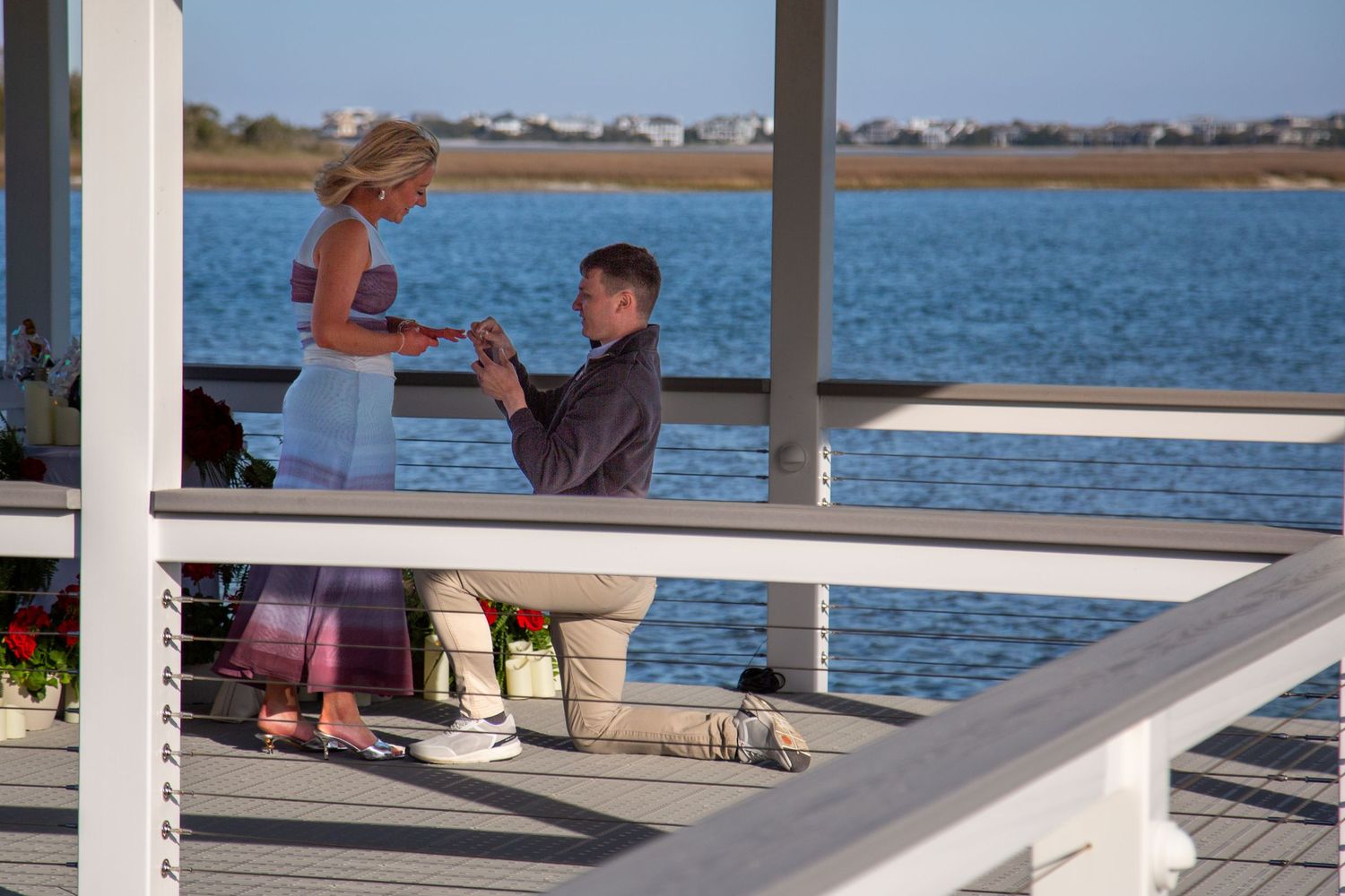Man proposes to woman on a waterfront dock, holding her hands on a sunny day near a calm blue bay.