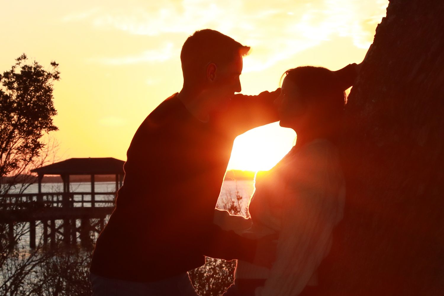 Romantic couple silhouetted against a golden sunset near a waterfront, sharing an intimate moment by a rocky wall.