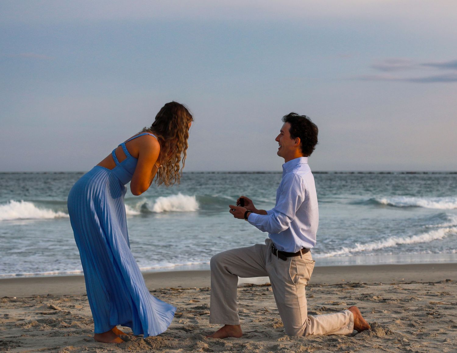 Man proposing on one knee to woman in blue dress on a sandy beach at sunset with ocean waves behind them.