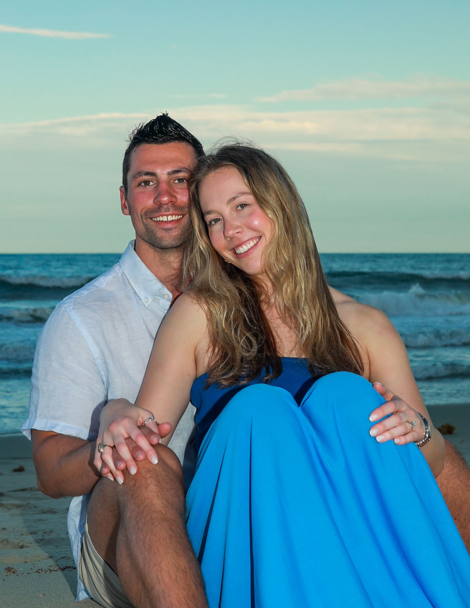 A smiling couple poses together on a sandy beach at sunset, the woman in a blue dress and man in a white shirt.