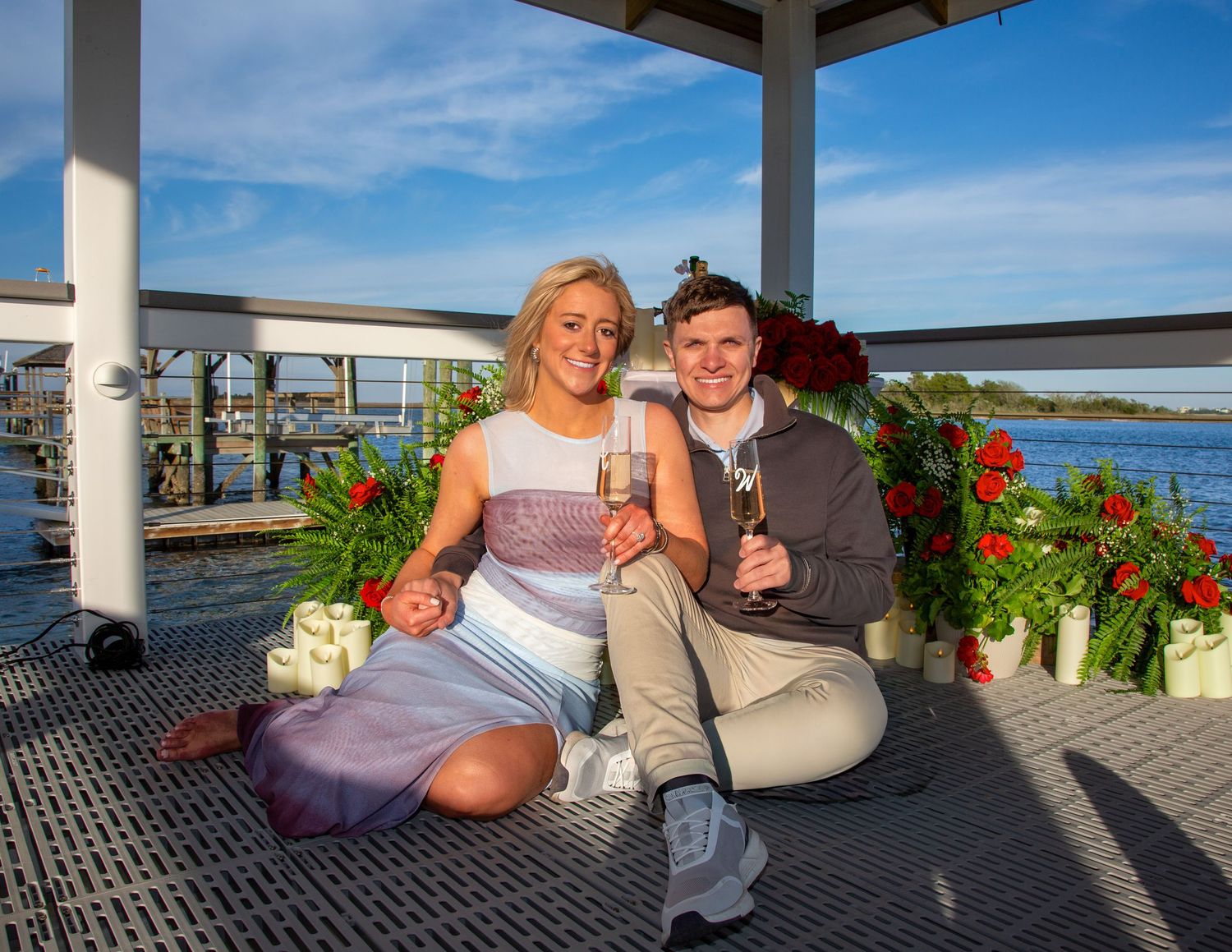 Couple sitting on a waterfront gazebo deck, holding champagne glasses, surrounded by red floral arrangements on a sunny day.