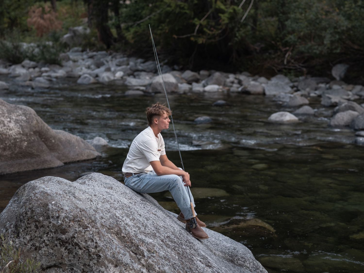 Young man sitting on a boulder beside a rocky river, holding a fishing rod in a serene outdoor setting.