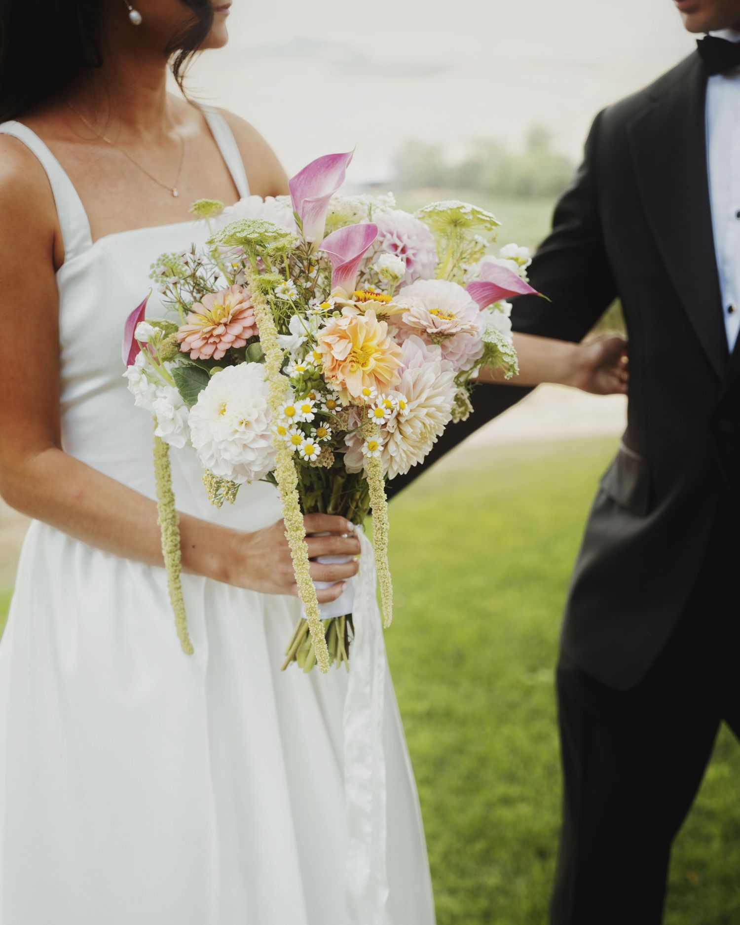 Bride holding a colorful wedding bouquet with pink calla lilies, dahlias, and wildflowers beside groom in black tuxedo.