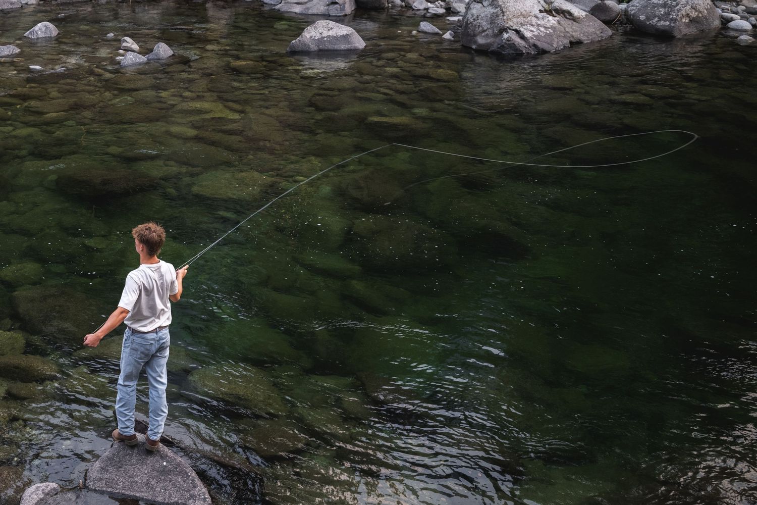 Young boy fly fishing on rocky riverbank with clear green water and mountains in background.