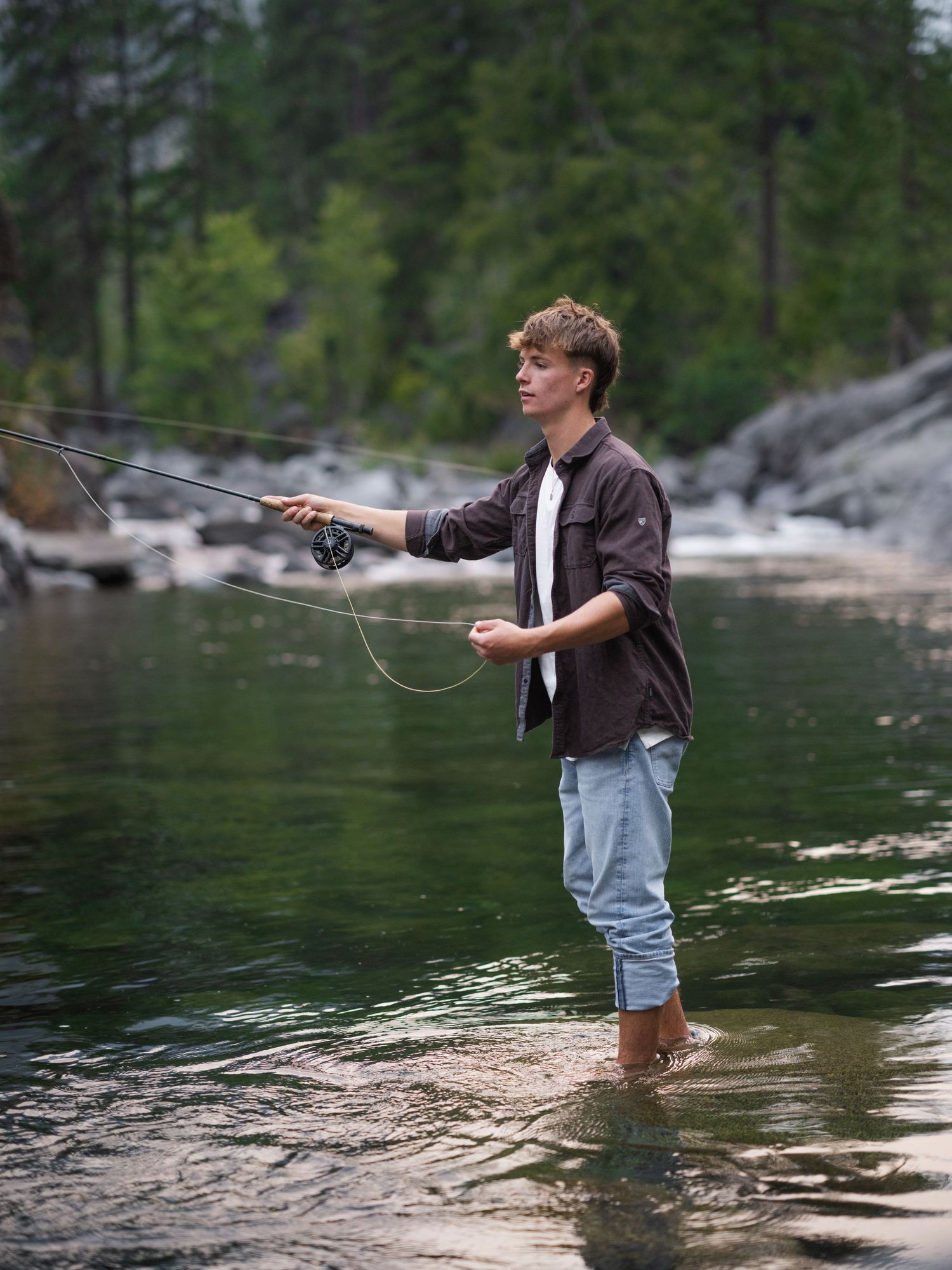 A young man fly fishing in a shallow river, wearing a brown shirt and light jeans, surrounded by forest.