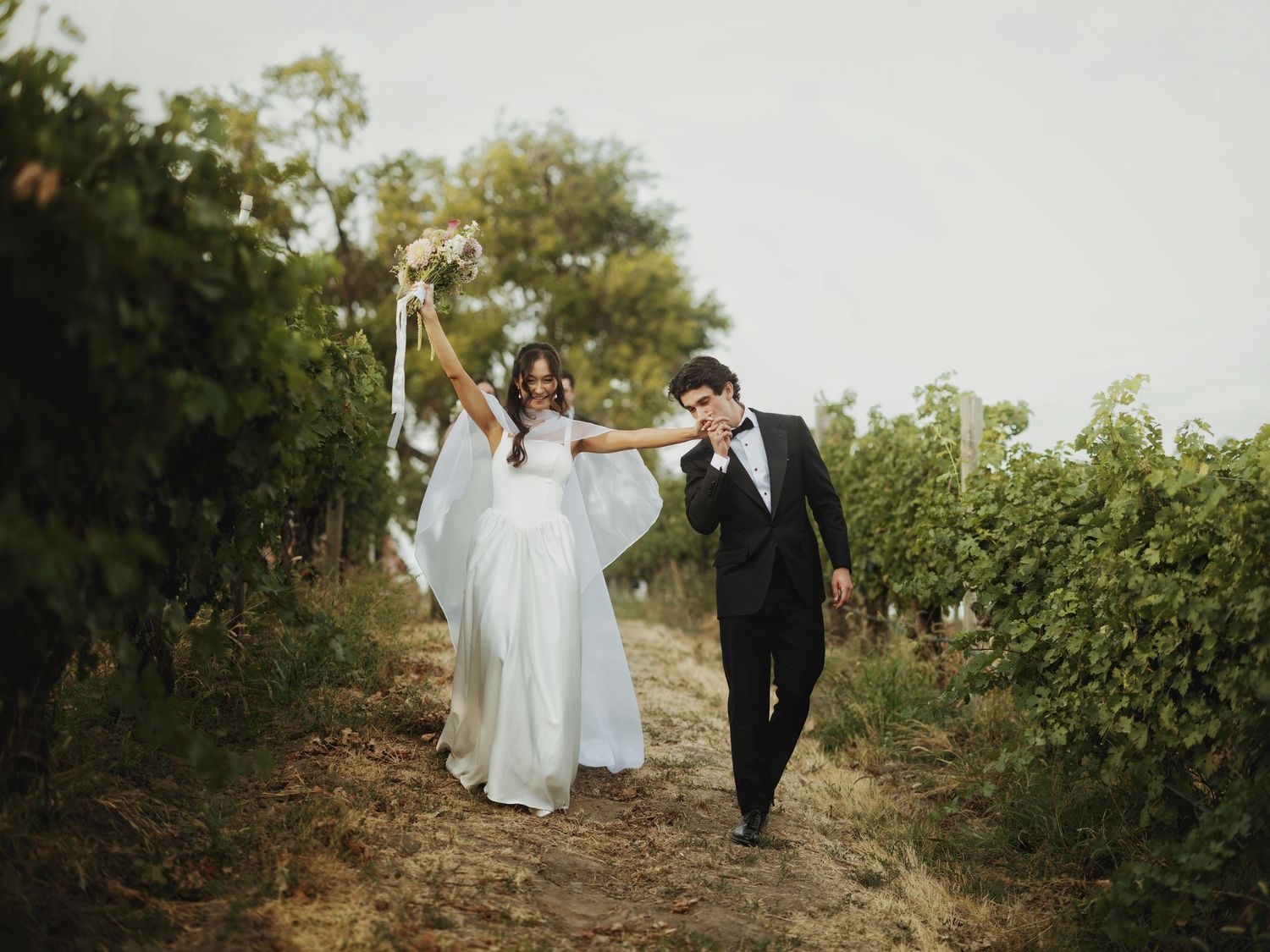 Bride and groom walking through a vineyard, bride holding bouquet aloft in a white gown, groom in black tuxedo.