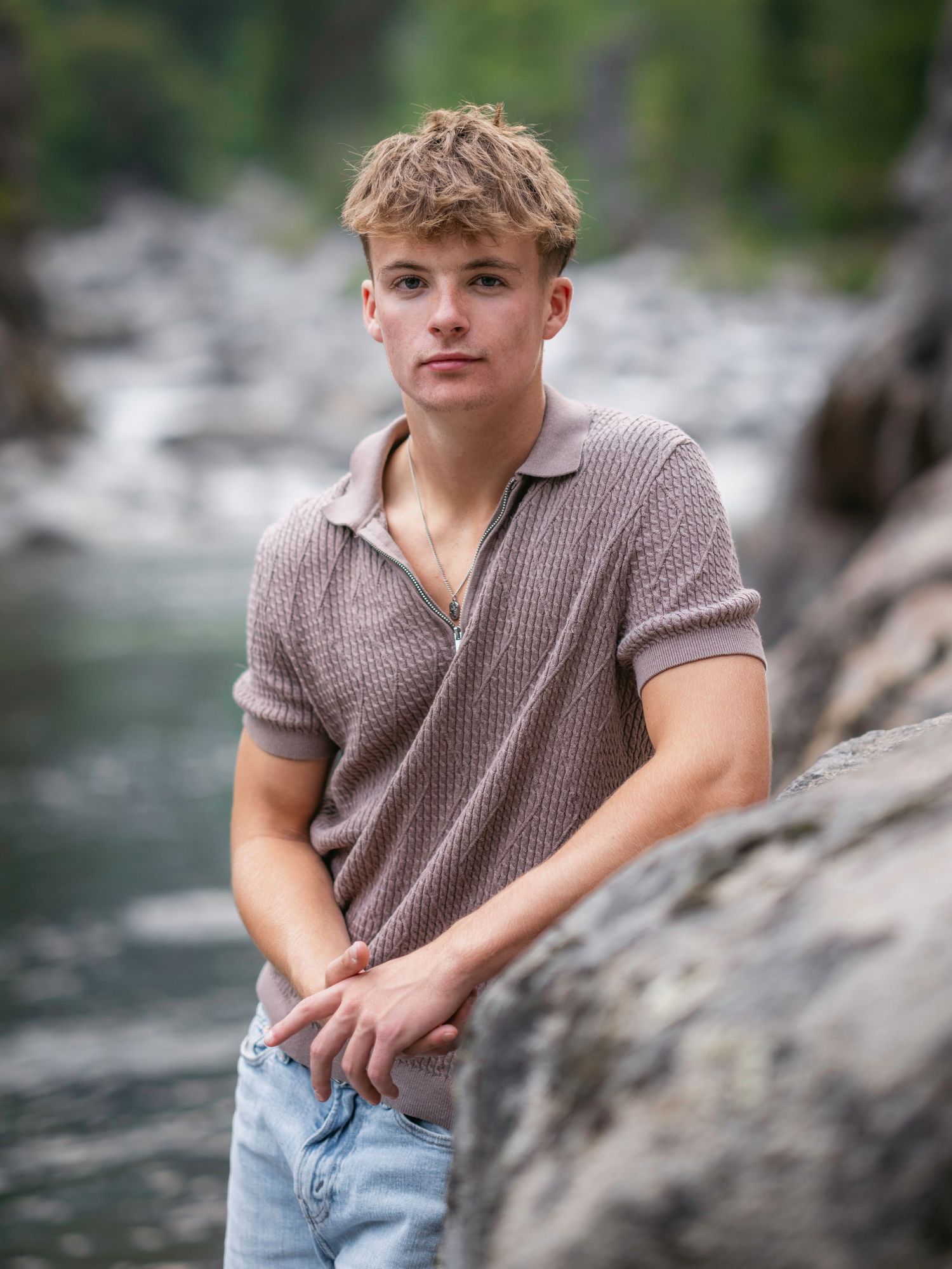 A young man in a brown knit polo and jeans poses on rocks near a river for senior portraits.