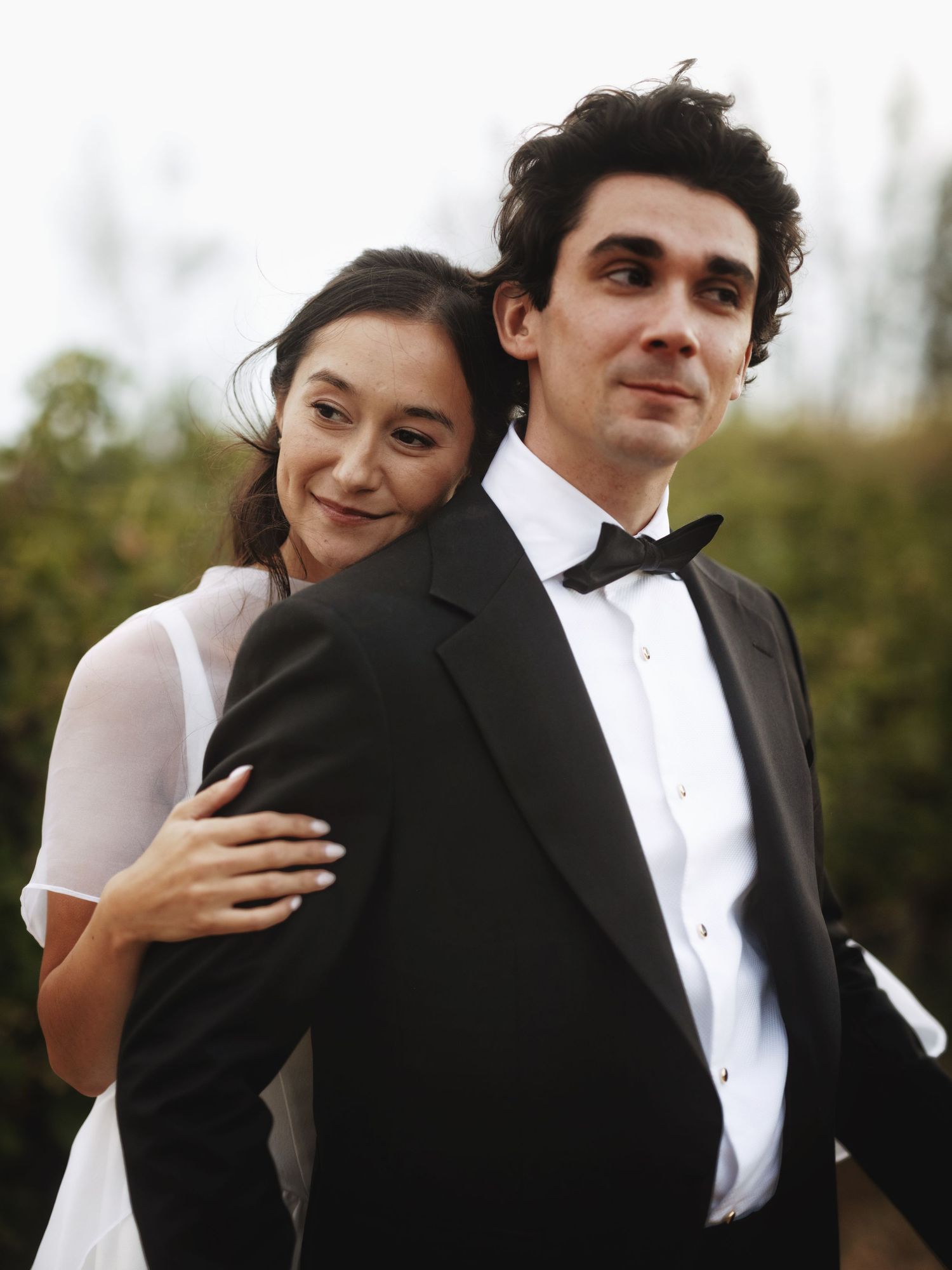Elegant couple in formal attire, woman in white dress embracing man in black tuxedo with bow tie outdoors.