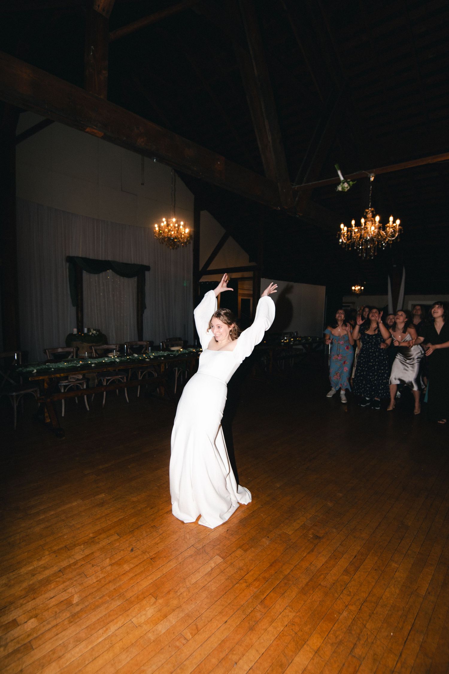 Bride in white gown tossing bouquet on dance floor at elegant wedding reception with chandeliers.