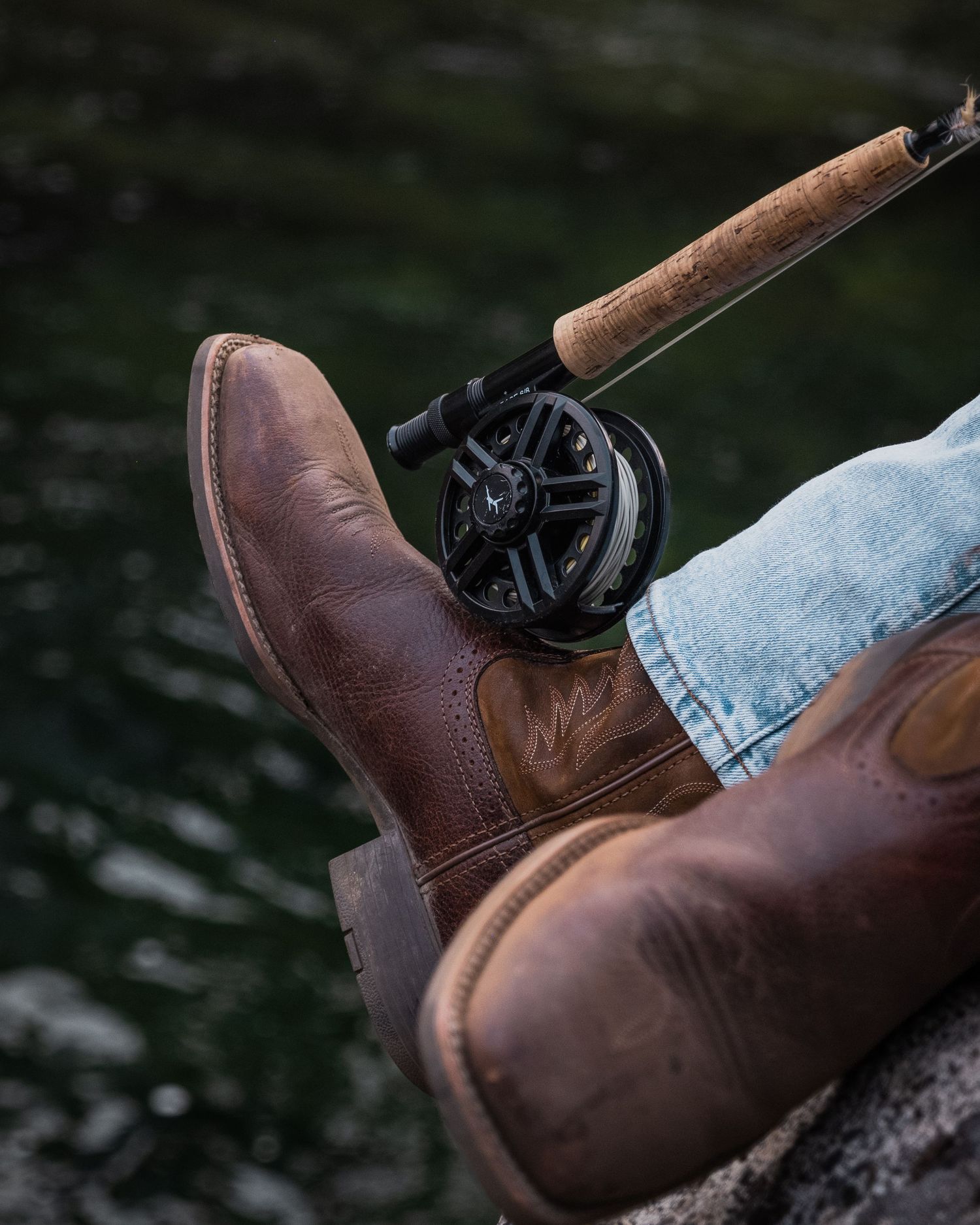 Person wearing brown cowboy boots and jeans resting by water while holding a fly fishing rod with black reel.
