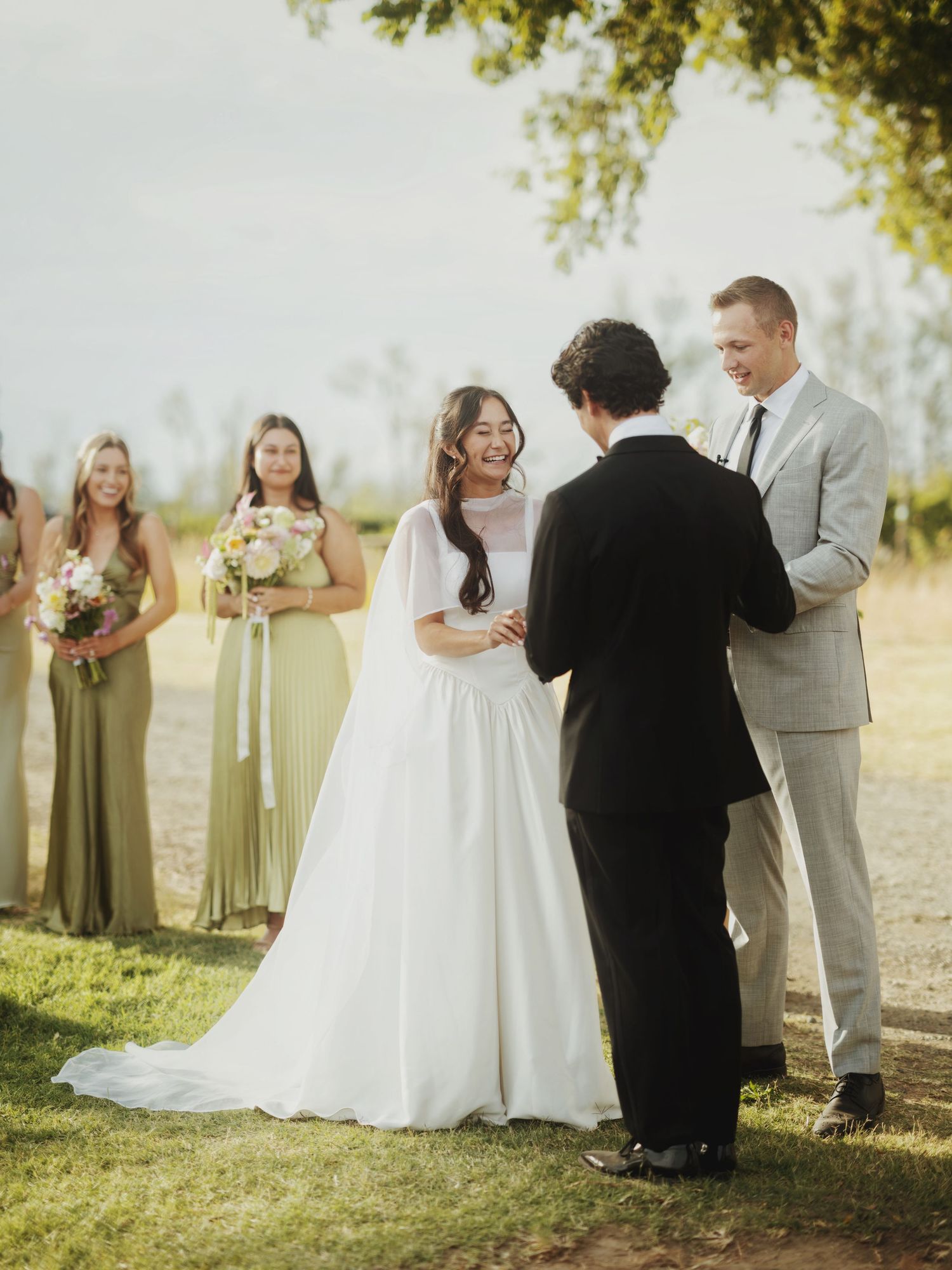 Bride and groom exchange vows outdoors with bridesmaids in sage green dresses and officiant during sunny wedding ceremony.
