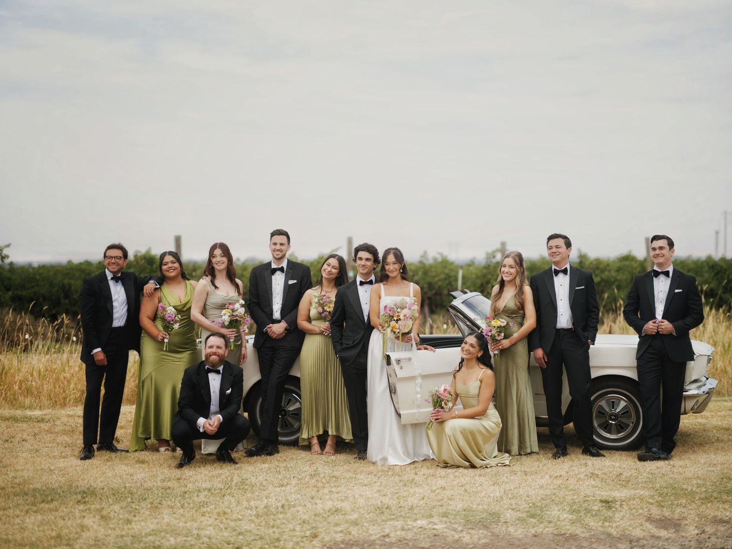 Wedding party poses outdoors in formal attire, bridesmaids in sage green gowns, groomsmen in tuxedos, with a vintage white car.