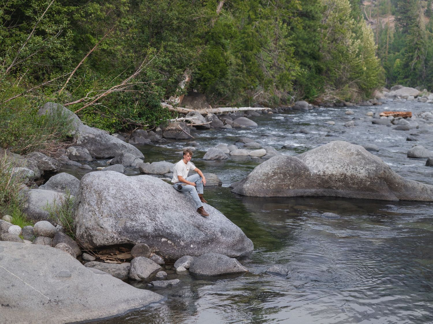 Person sitting alone on large boulders beside a scenic rocky mountain river surrounded by trees.