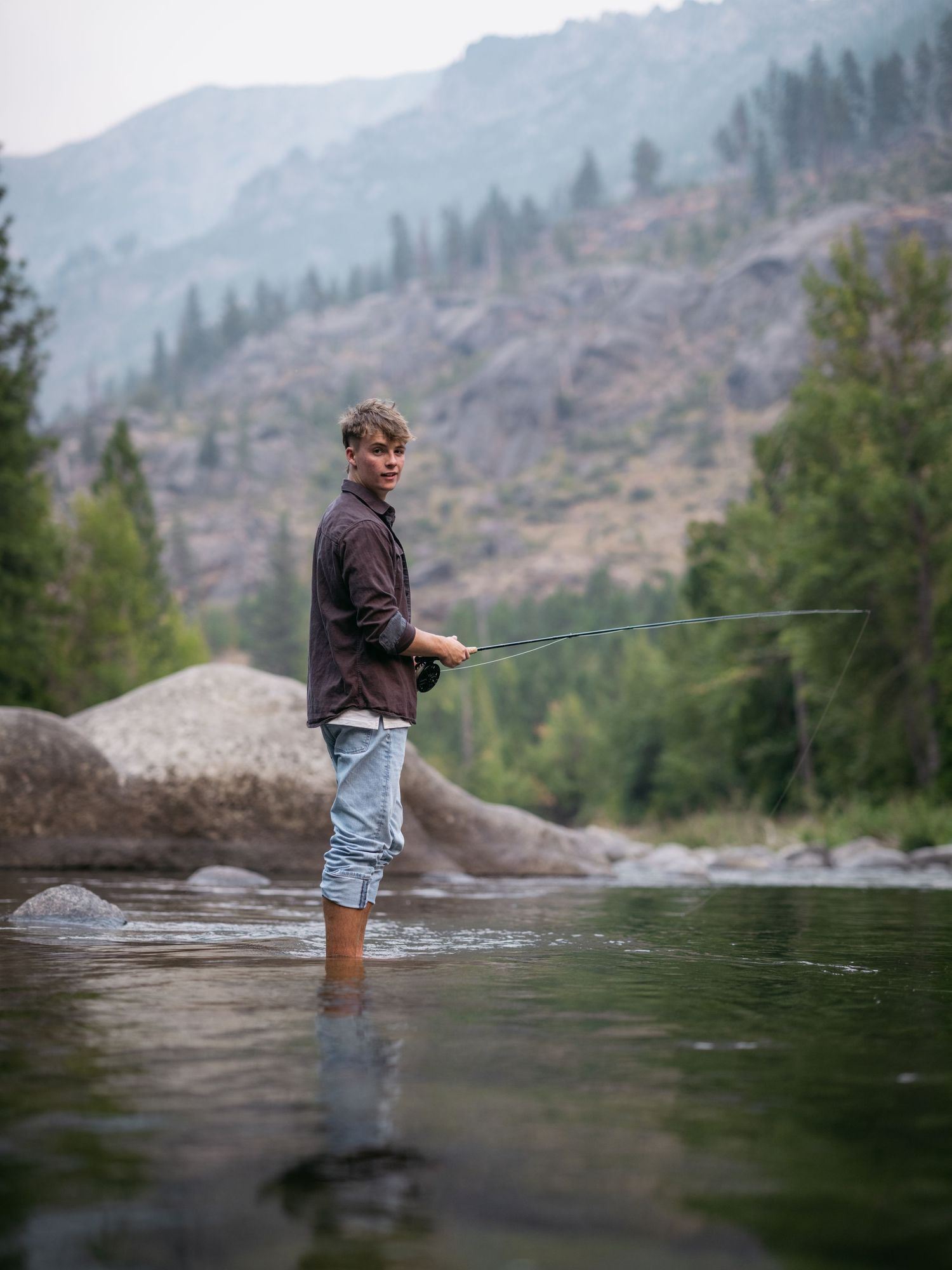 Teen boy fly fishing while standing in a shallow mountain river, holding a fishing rod.