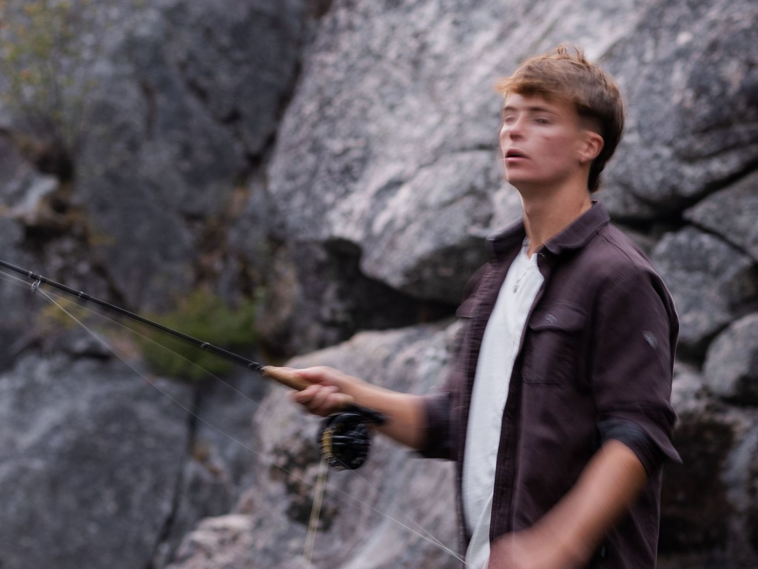 Young man fly fishing outdoors, holding a fishing rod near rocky cliffs in natural light.