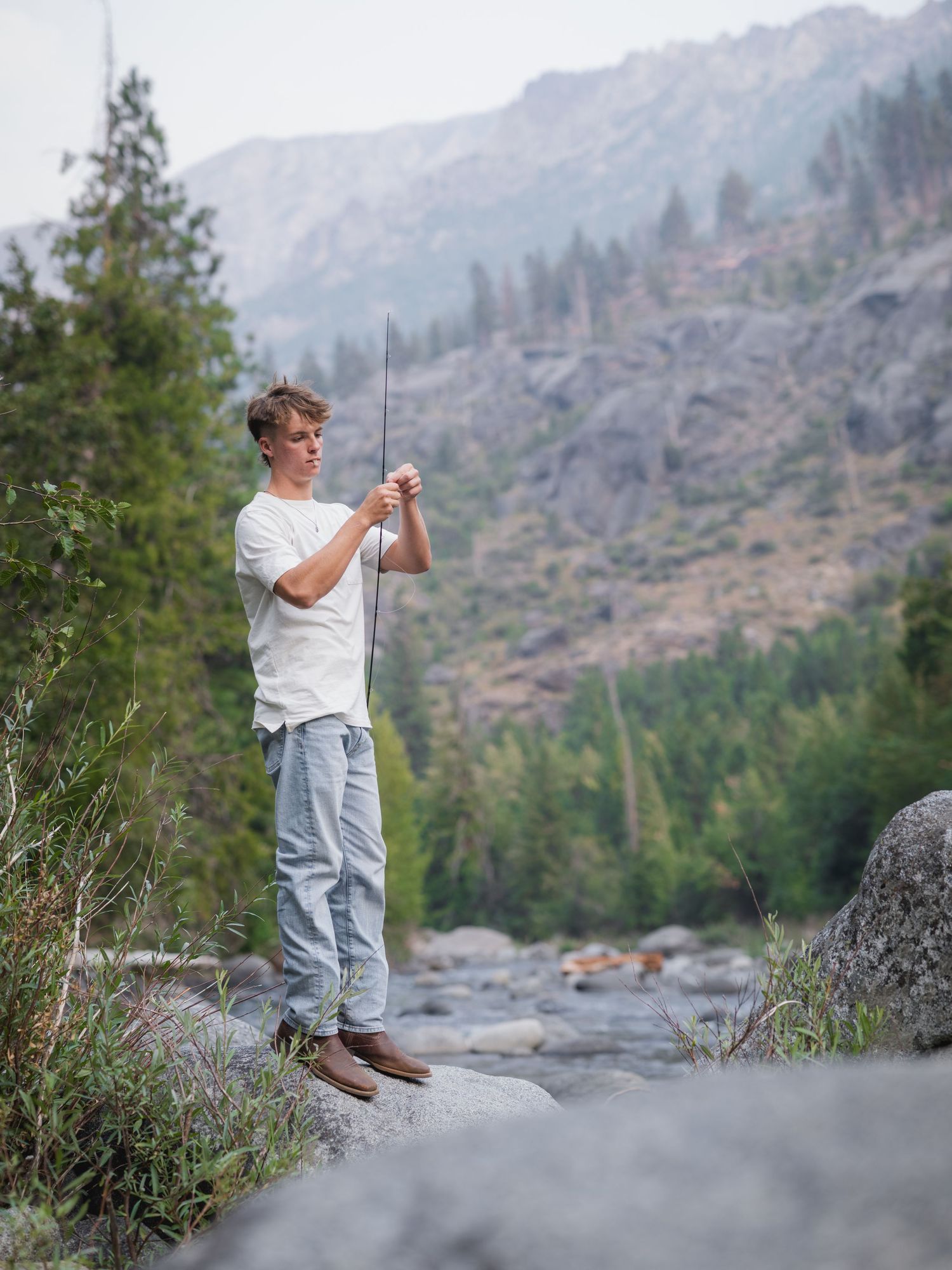 Young man fly fishing on rocky riverbank with mountain forest backdrop.