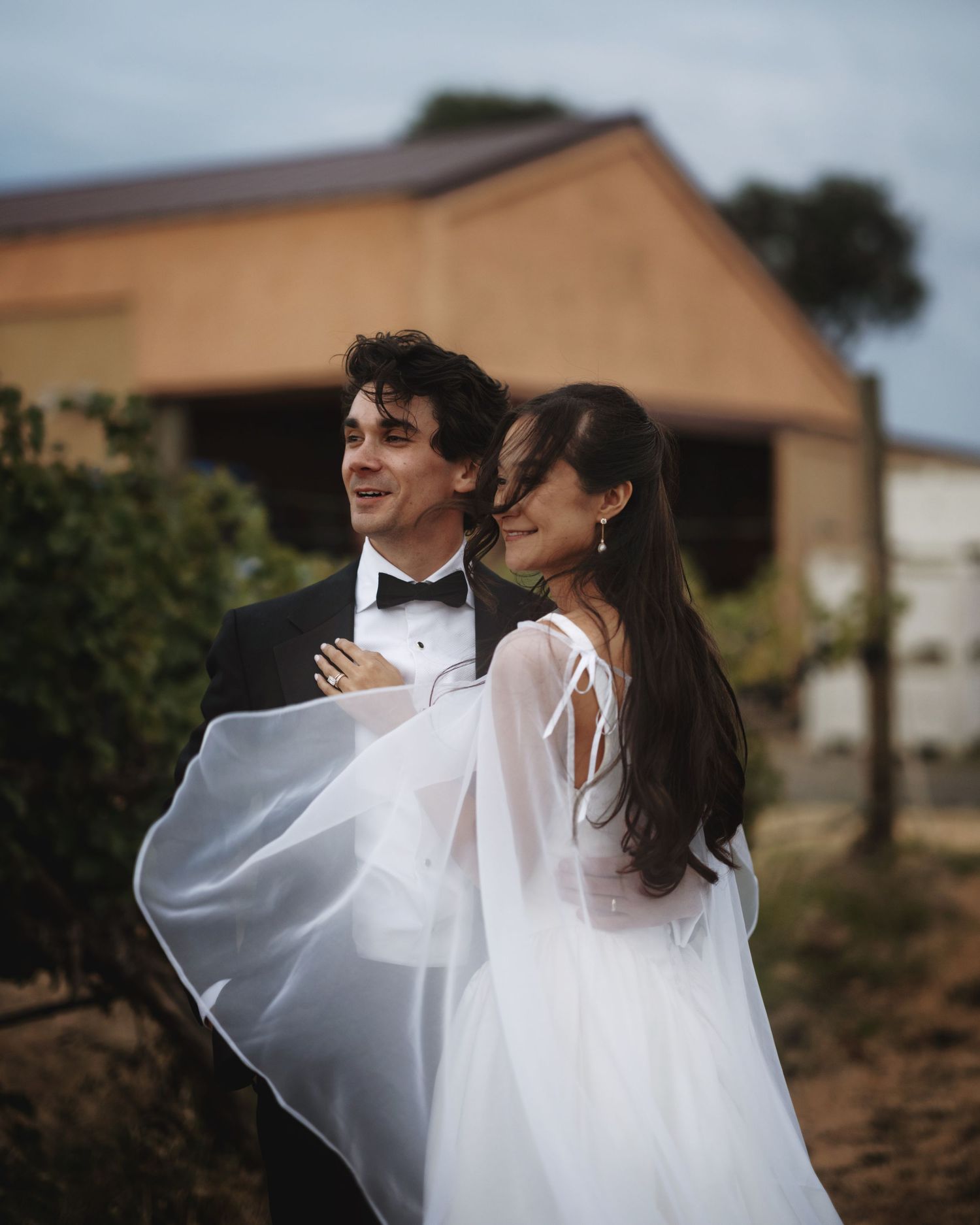 A couple in wedding attire smiles outdoors, bride in white gown with flowing cape, groom in black tuxedo.