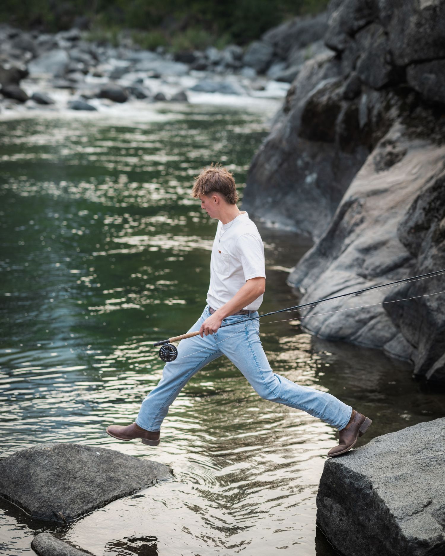 Young man stepping across rocks by a river while fly fishing, wearing jeans and a white shirt.