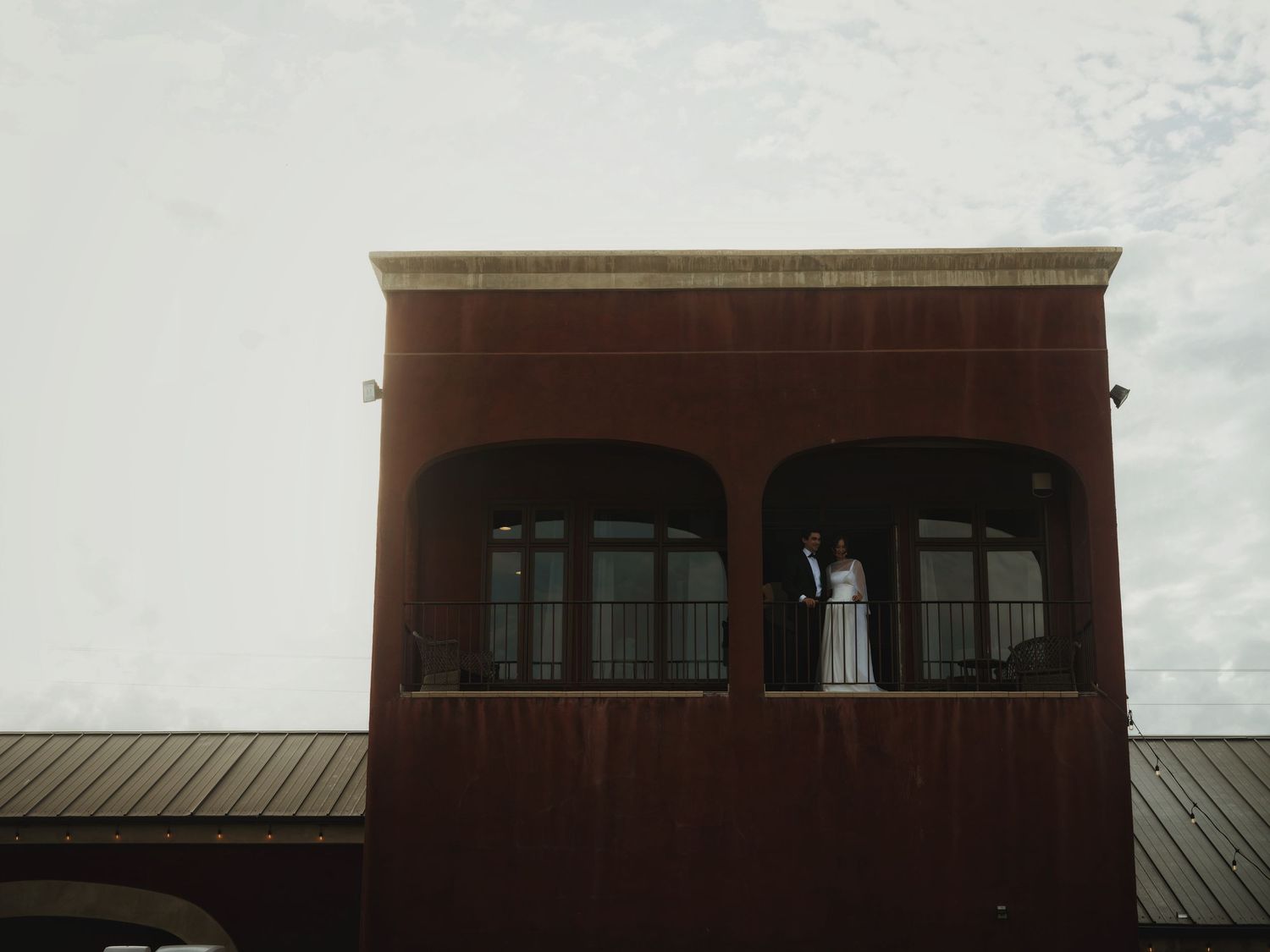 Bride and groom standing on a red brick building balcony with arched windows on their wedding day.