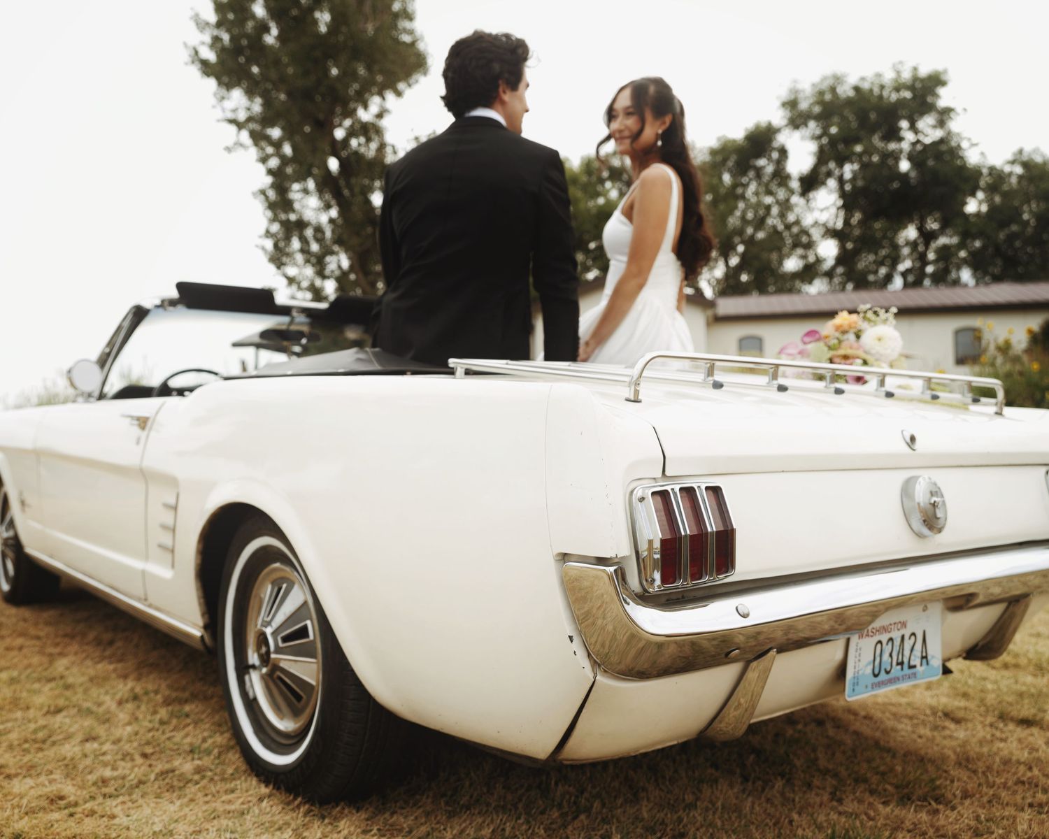 Bride and groom standing beside a classic white Ford Mustang convertible at an outdoor wedding celebration.