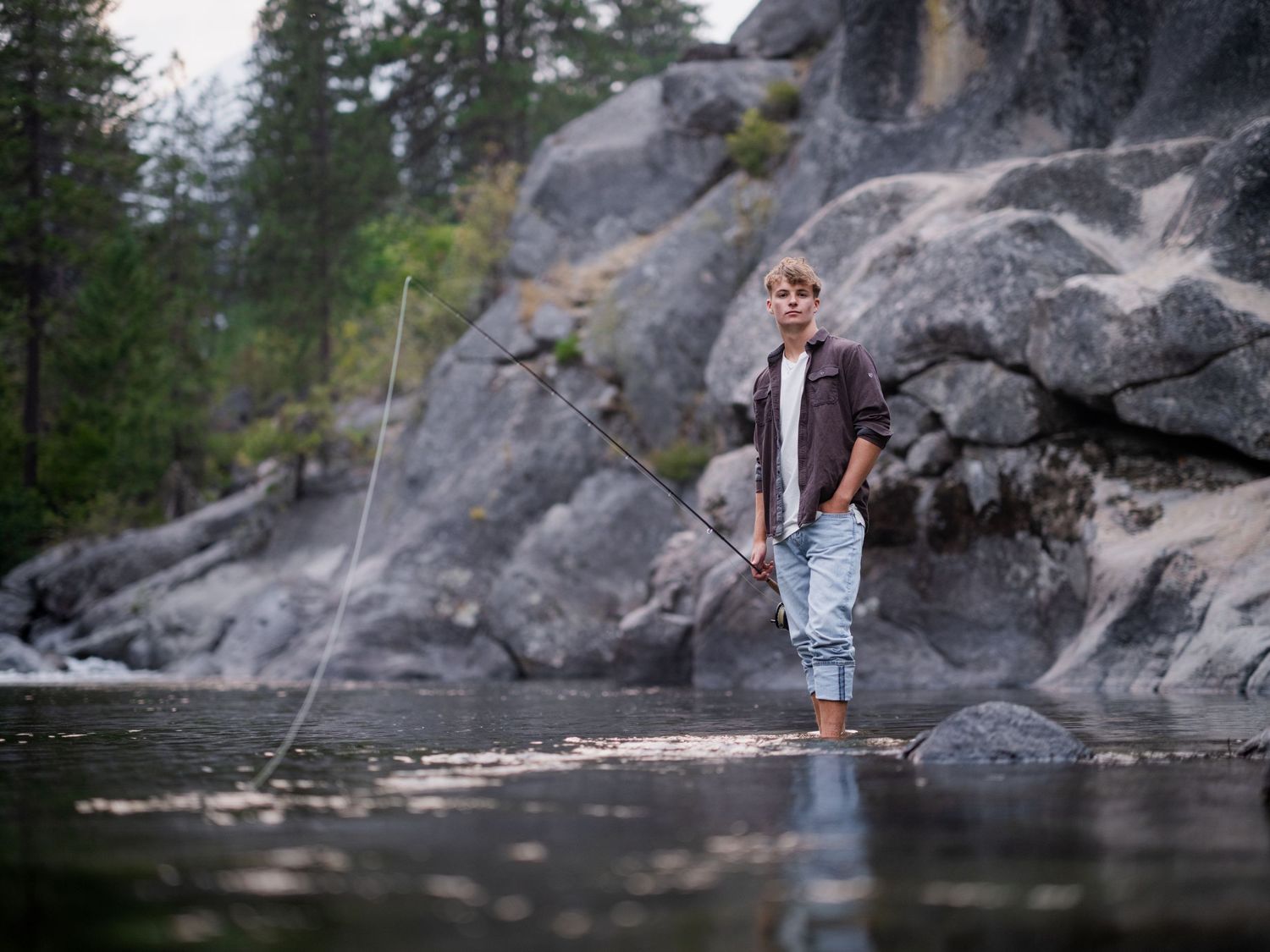 A young boy stands in a shallow river holding a fishing rod, surrounded by rocky cliffs and pine trees.