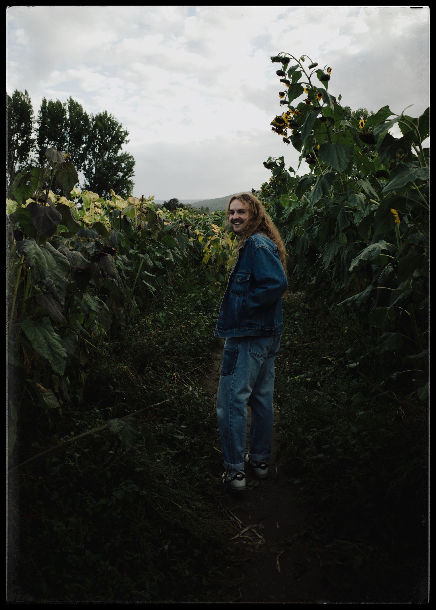 Person with long hair smiling in a sunflower field path, wearing a denim jacket and jeans on a cloudy day.