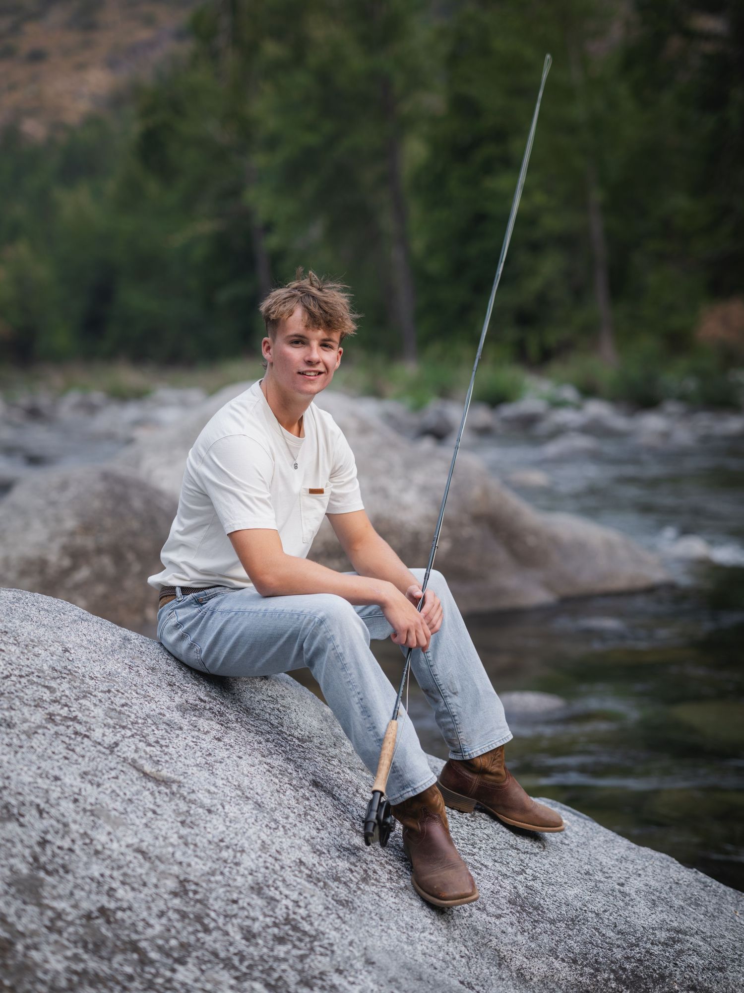 Young man sitting on river rocks with a fishing rod, wearing jeans and boots, with forest backdrop.