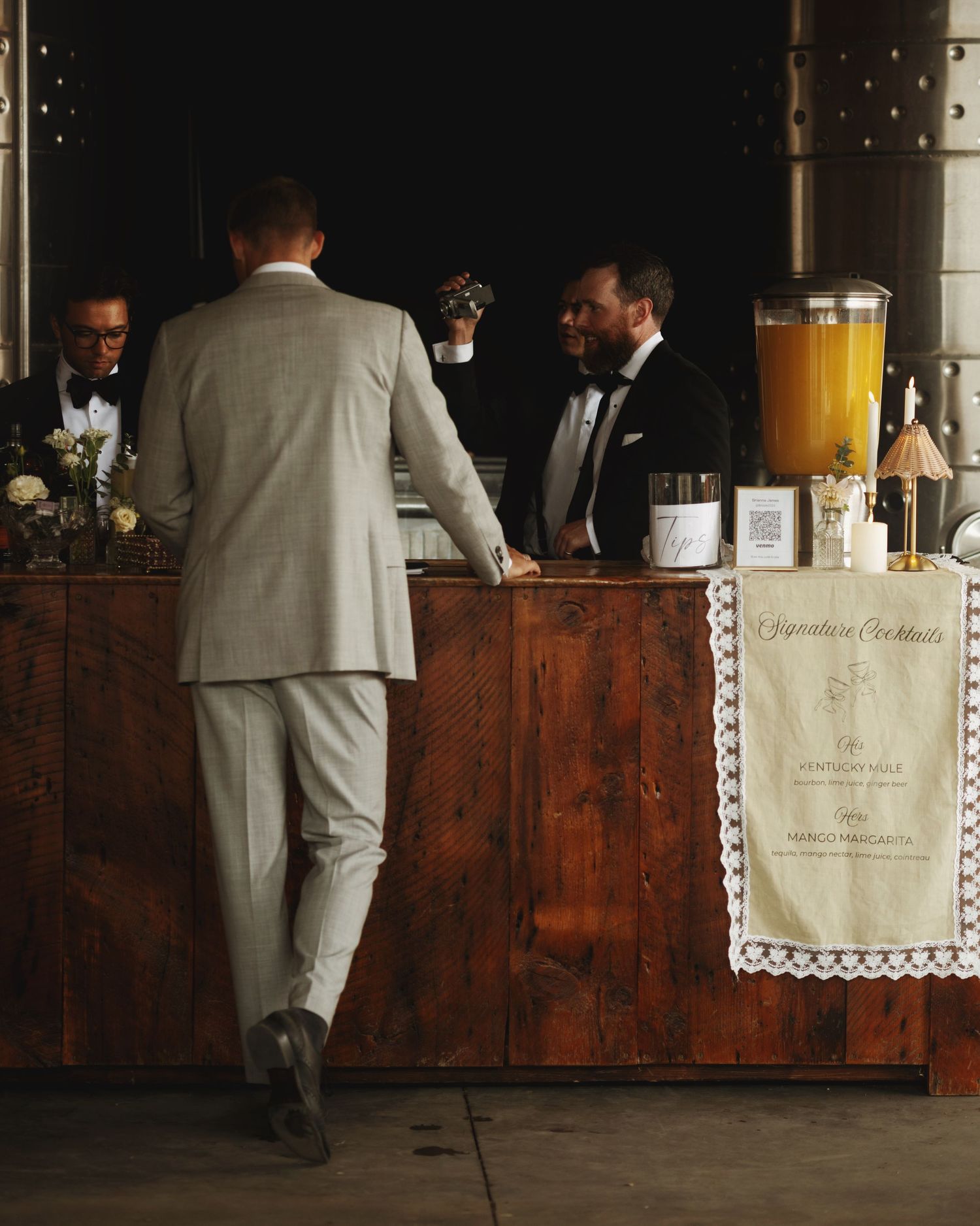 Elegant wedding bar setup with bartender in tuxedo serving signature cocktails from rustic wooden bar.