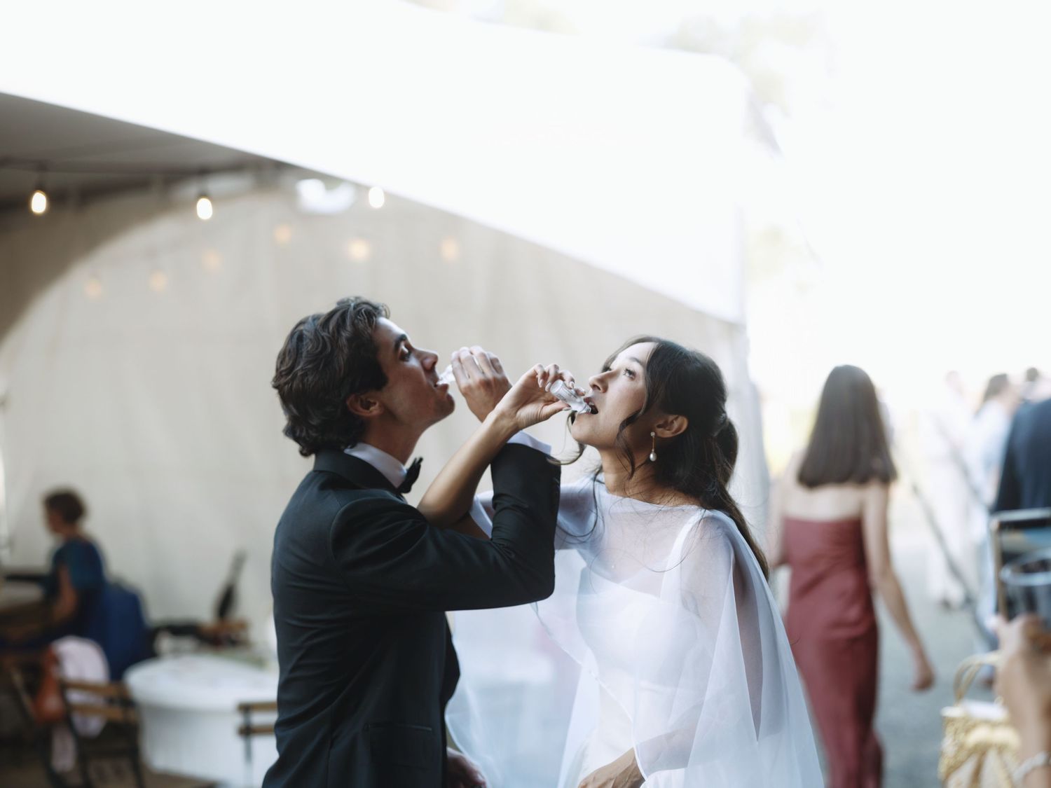 Bride and groom sharing a celebratory drink together outdoors at their wedding reception under string lights.