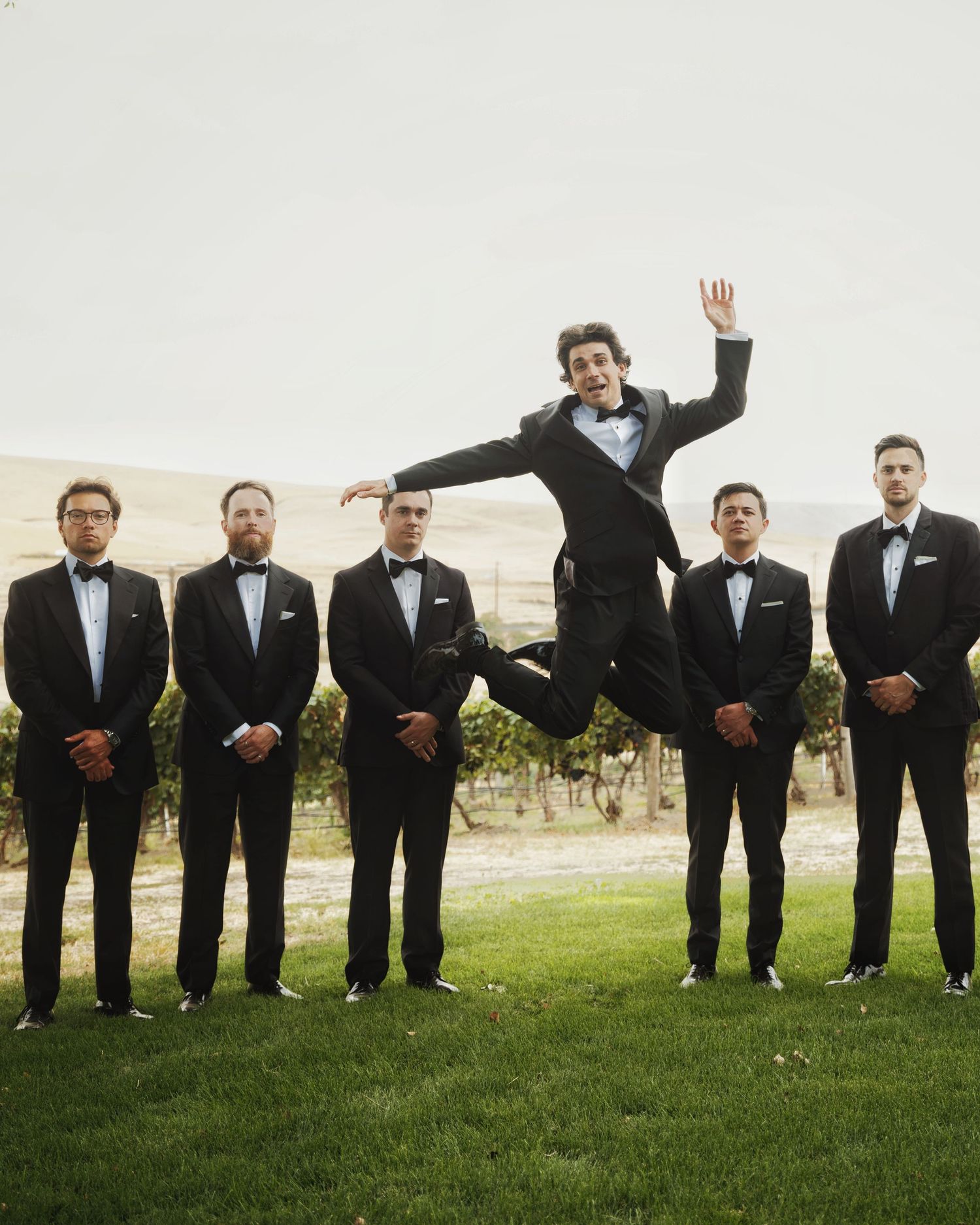 Groom jumping joyfully in the air surrounded by groomsmen in black tuxedos at an outdoor vineyard wedding.