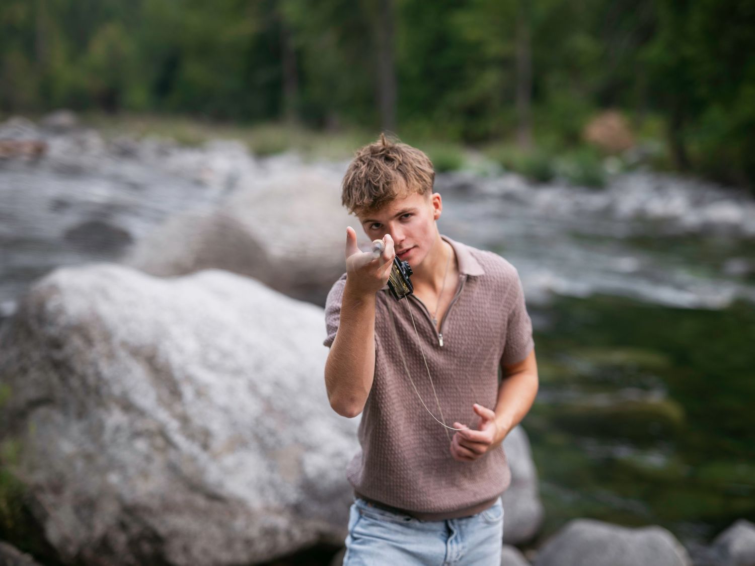 Young man holding a camera near a rocky river surrounded by lush green trees outdoors.