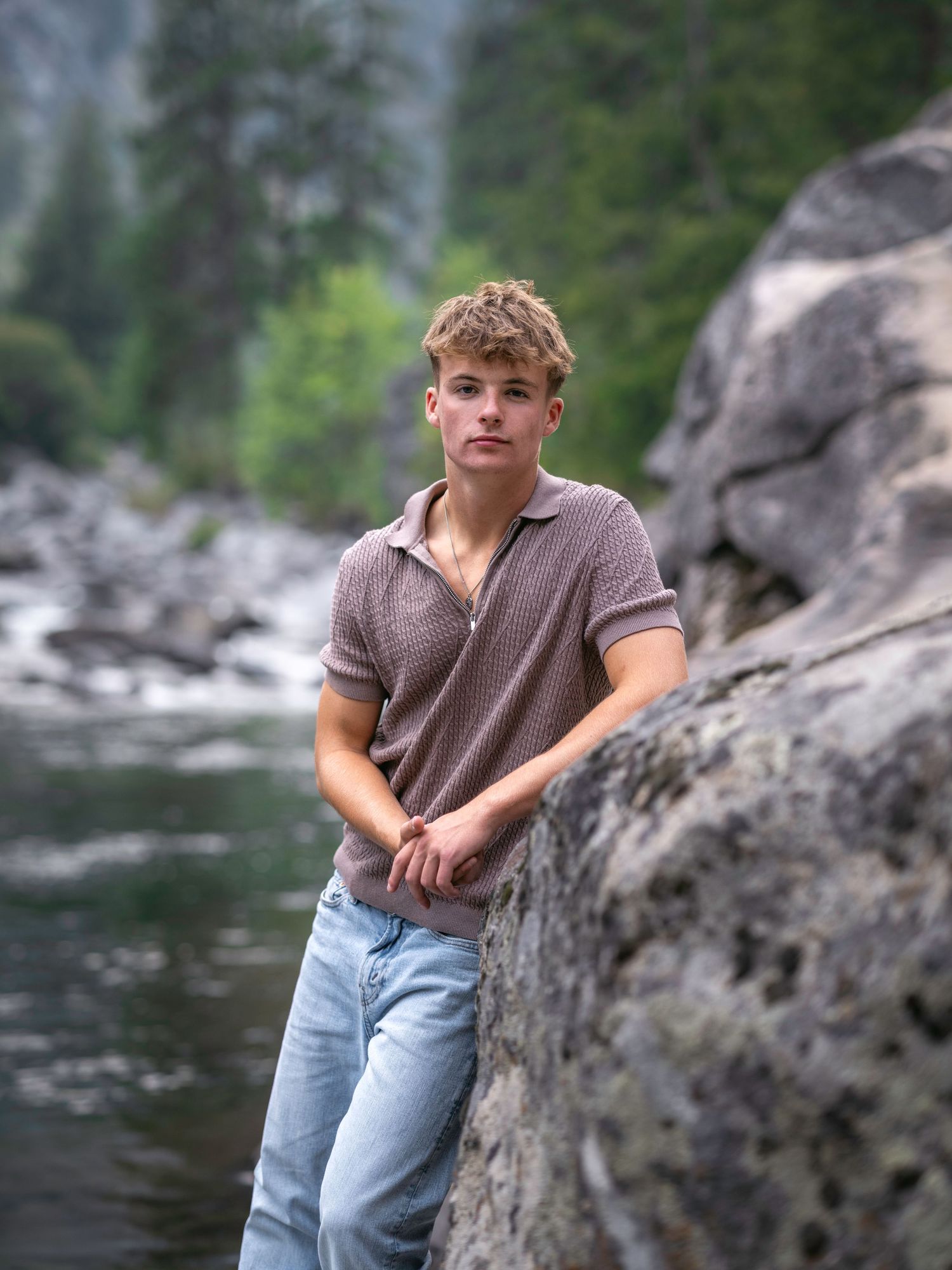 Young man in pink polo and jeans leaning on rocks by a river in a forested mountain setting.