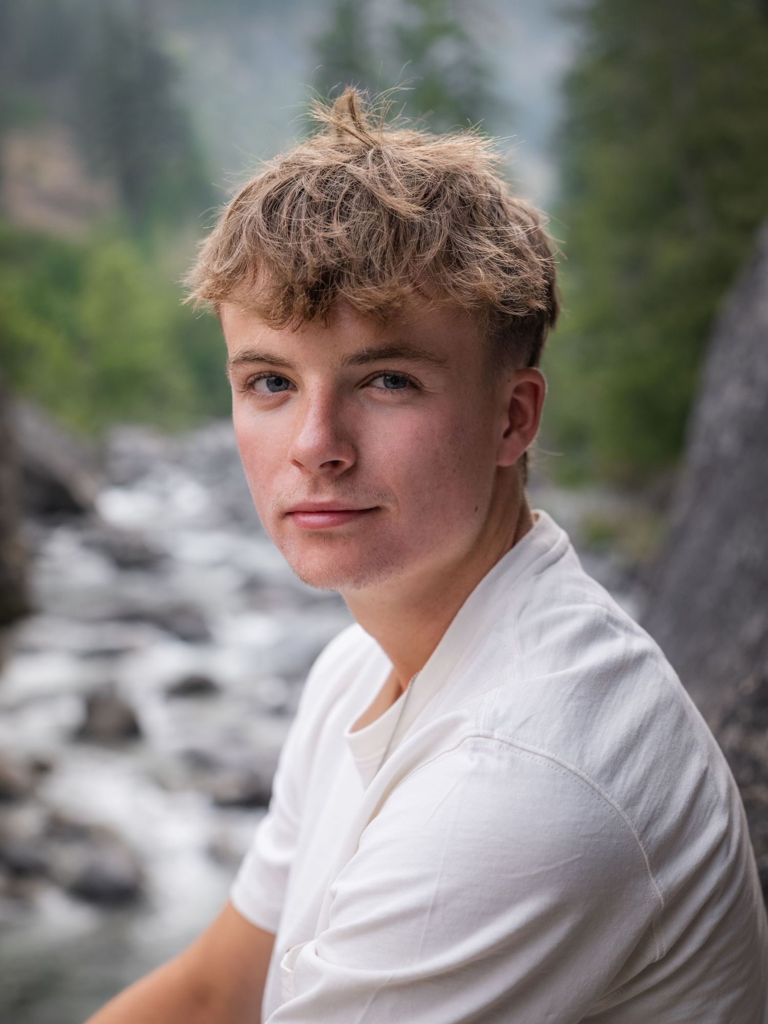 Young man with blonde hair in white t-shirt poses by a rocky mountain river in a senior portrait.