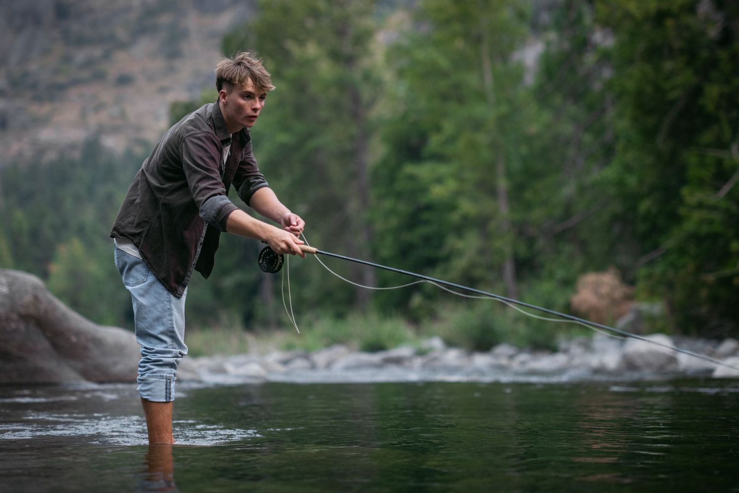 Man fly fishing in a scenic mountain river, casting a line while standing in shallow water.
